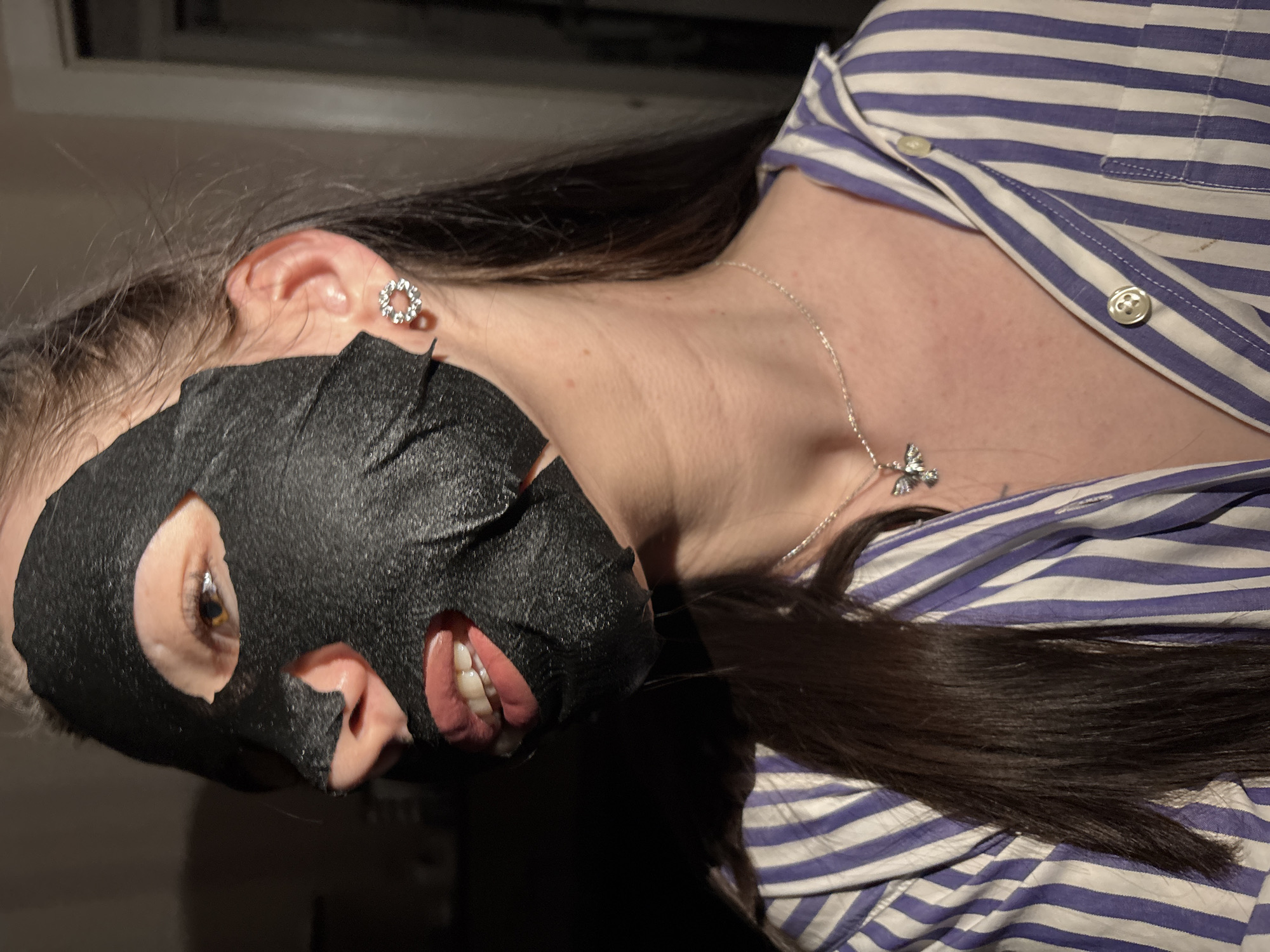A woman with dark brown hair is wearing a black face mask. She has a silver necklace with a butterfly pendant and a silver stud earring. She is wearing a blue and white striped button-down shirt. The location of Perm, Russia is not determinable from the provided image.