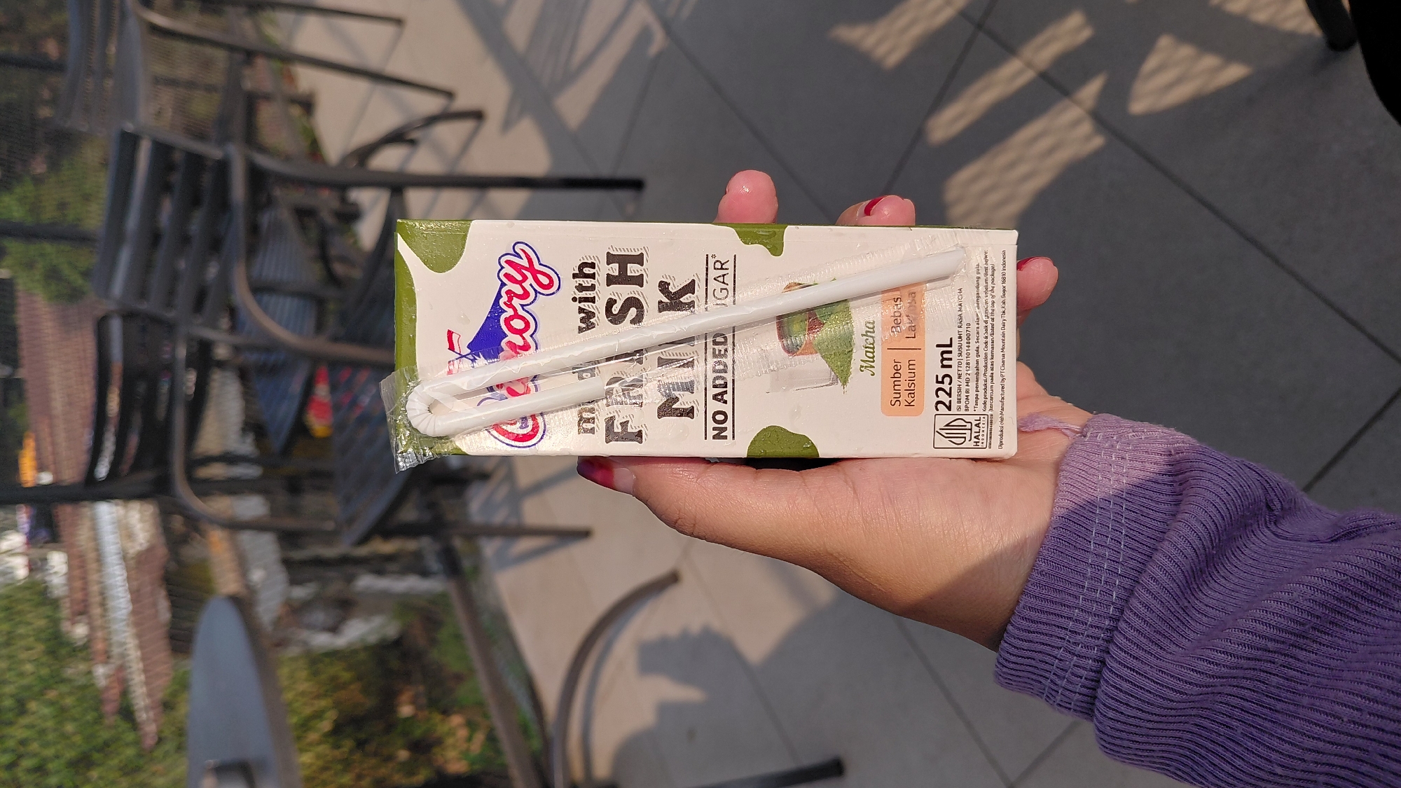 A hand wearing a purple long-sleeved shirt holds a 225 mL carton of "Cemory Milk With Fresh Milk No Added Sugar Matcha" flavored UHT milk. The carton has a blue and red logo with the text "Cemory" in cursive script. There are two white straws inserted through a plastic wrapper on the front of the carton. Text on the carton includes "Sumber Kalsium" (Calcium Source) and "Bebas Laktosa" (Lactose-Free). Below this, there is a "HALAL" certification mark and BPOM registration number. The text "Diproduksi oleh: Manufactured by PT. Ciarmoy Mountain Dairy Tbk. Kab. Bogor 16810 Indonesia" indicates the manufacturer's location. The background shows outdoor seating with dark metal chairs and tables, suggesting a cafe or restaurant setting. The scene is illuminated by natural sunlight, casting shadows. While the image is taken in Karanglewas, Indonesia, there are no specific visual cues in the image to confirm this exact location.