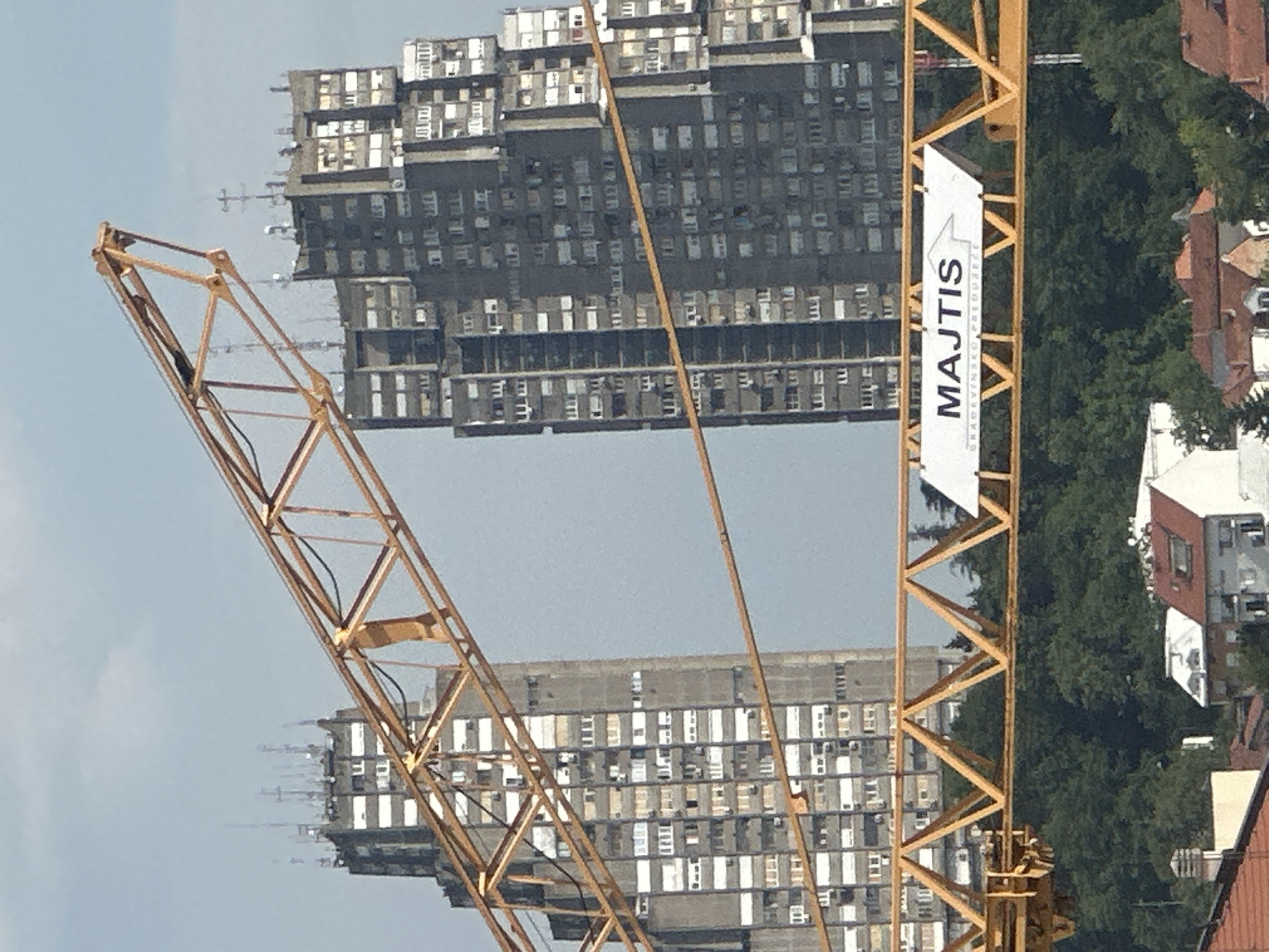 The image shows a construction crane in Belgrade, Serbia, with two large apartment buildings in the background. The crane is yellow and made of metal lattice. A white banner with the text "MAJTIS" is attached to the crane. The apartment buildings are tall and appear to be old, with many windows. The sky is blue with some clouds. The image is taken from a low angle, looking up at the buildings and the crane. The overall impression is one of ongoing construction and urban development.
The apartment buildings are characteristic of Brutalist architecture, common in former Yugoslavia. They are monolithic and imposing, with repetitive window patterns. The crane suggests that either these buildings are under renovation or new construction is taking place in the vicinity. The banner on the crane indicates a construction company, "MAJTIS", which is a Serbian company specializing in building construction and reconstruction. The presence of trees and other buildings suggests an urban setting. The time of day appears to be daytime, possibly late morning or early afternoon, given the lighting. The weather seems to be clear and sunny.