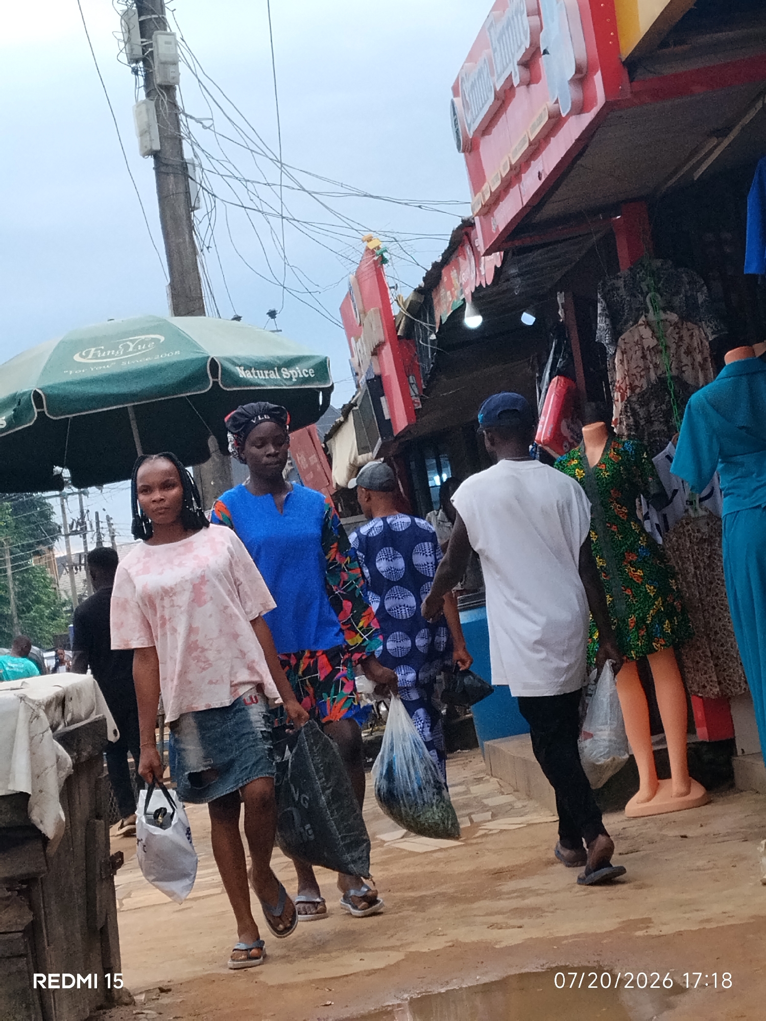 The image captures a street scene in Umuode, Nigeria. A group of people are walking along a dusty path in front of several market stalls. In the foreground, a young woman in a pink tie-dye shirt and a distressed denim skirt walks towards the viewer, carrying a white plastic bag. To her left, another young woman in a blue top with a patterned skirt also carries a large, dark plastic bag. Behind them, a man in a white sleeveless shirt and black trousers walks away from the camera. In the background, market stalls display clothing and other goods. A green umbrella with "Fung Yue Natural Spice" printed on it is visible. Power lines crisscross overhead. The ground is uneven and appears to have patches of mud. The timestamp "07/20/2026 17:18" is present in the bottom right corner, along with "REDMI 15" in the bottom left.