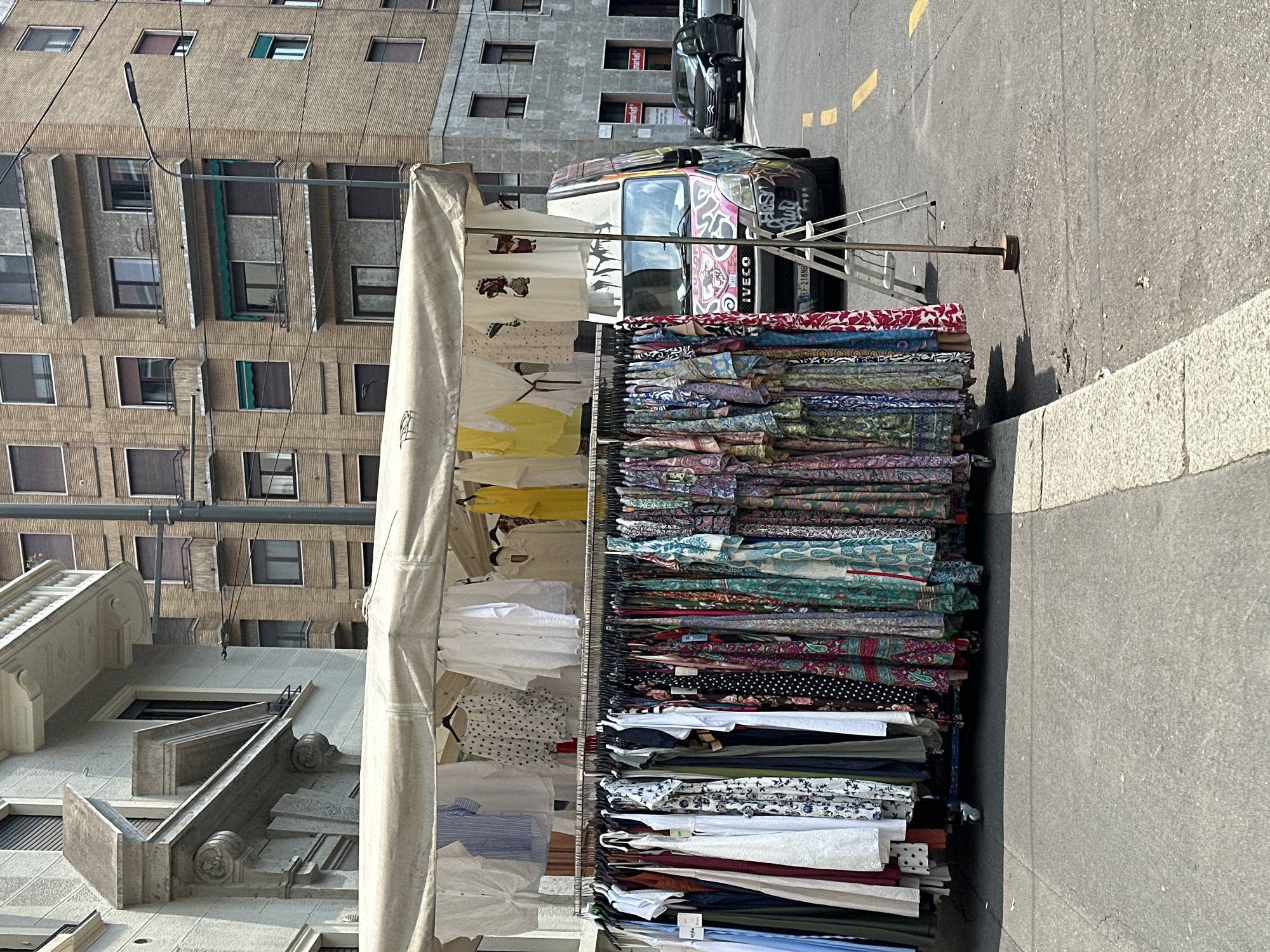An outdoor clothing stall is set up on a street in Milan, Italy, featuring a variety of dresses and tops displayed on racks. A white canopy covers the stall, and a colorful, graffiti-adorned Iveco van is parked behind it. In the background, a brick building with multiple windows and balconies looms over the scene, typical of Milanese architecture. A black car and a silver car are visible on the street. The image captures a sunny day, with shadows cast by the stall and street furniture. The foreground shows the pavement and a concrete curb.