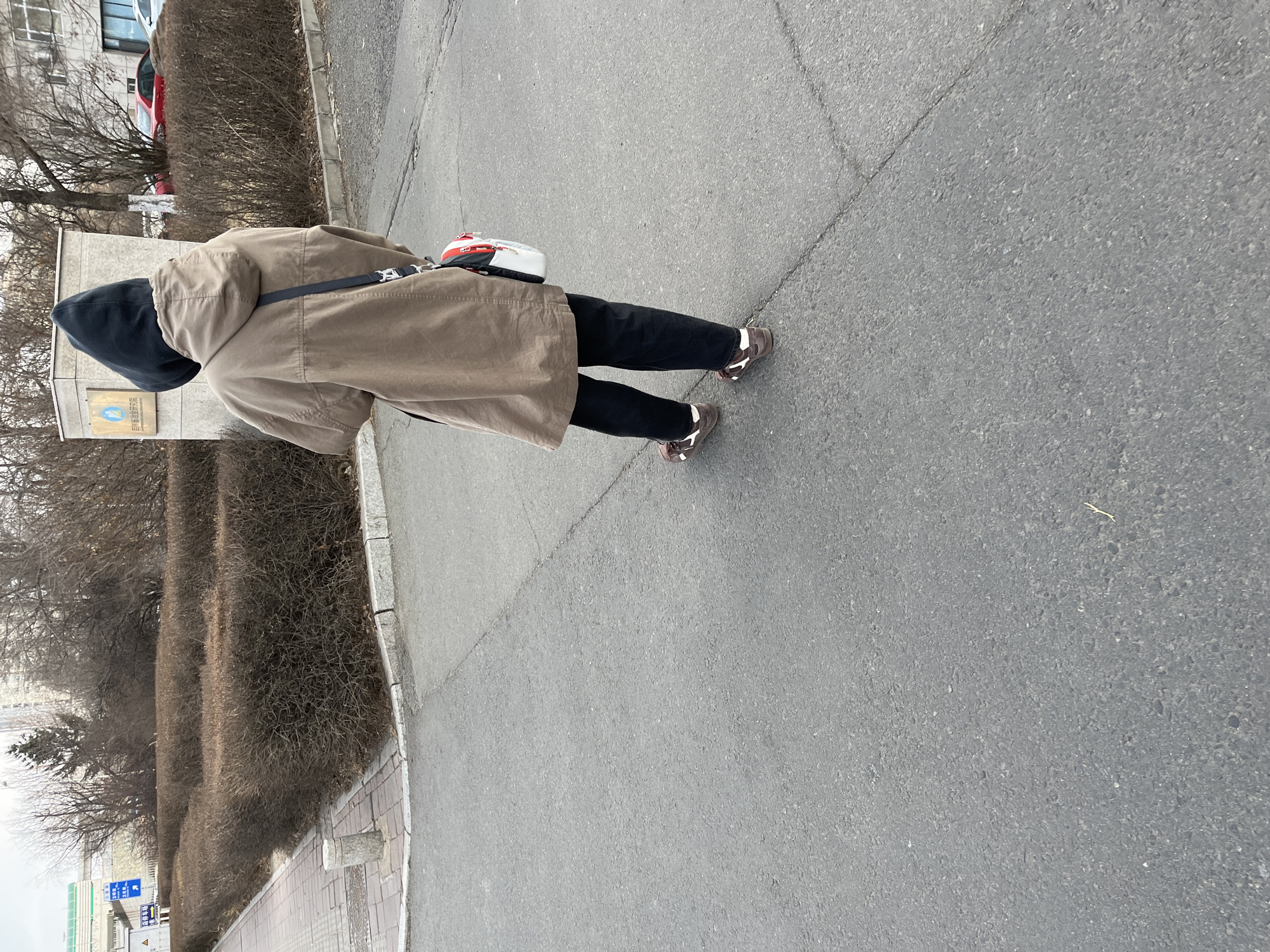 A person wearing a hooded sweatshirt and khaki coat is walking on an asphalt sidewalk in Changchun City, China. The person has black pants and brown and white shoes. They are carrying a white and red bag over their shoulder. There are bushes and trees alongside the sidewalk. In the background, there is a building with Chinese writing on a plaque and a glimpse of a red car.