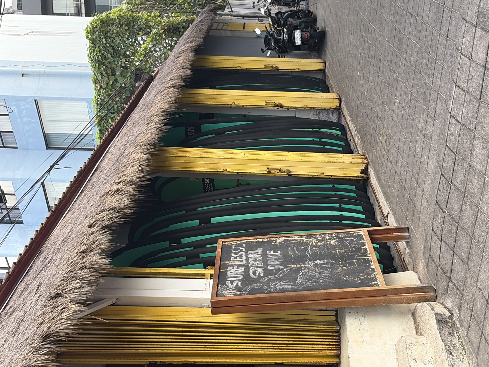 The image shows a low-angle shot of a surf shop entrance with a thatched roof. The shop appears to be located in Kuta, Indonesia, indicated by a partial view of a blue building with horizontal windows and a tiled exterior wall. Stacked vertically inside the open entrance are numerous surfboards, mostly dark with turquoise accents. Yellow metal shutters are visible on either side of the surfboard display.
In the foreground, a rustic chalkboard sign is propped up. White chalk writing on the board reads "SURF LESSONS SPECIAL PRICE". Behind the sign and extending into the background, more surfboards are neatly arranged. Above the thatched roof, a glimpse of green foliage from an overgrown plant is visible, along with some power lines. In the upper right portion of the image, two motorcycles are parked on a tiled walkway, suggesting a busy street or public area. The overall impression is a commercial setting, likely a rental or lesson provider catering to surfers in a tropical location. The lighting suggests it could be daytime, but the weather is not definitively clear.