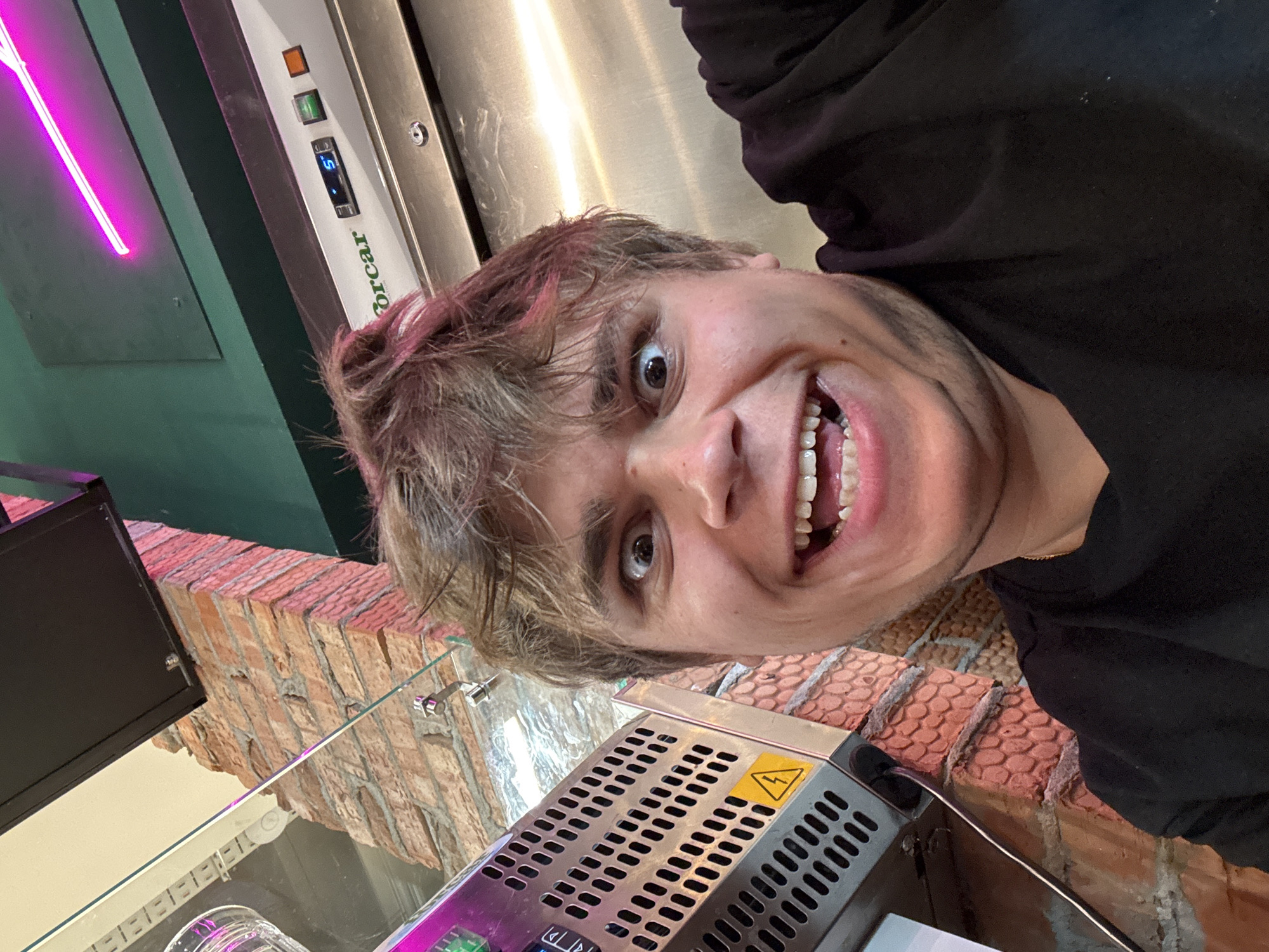 The media file captures a close-up, rotated selfie of a young person with tousled, light brown hair, possibly with hints of pink dye. The subject is looking directly at the camera with a wide, open-mouthed smile, revealing their teeth and tongue. Their eyes are wide open, conveying a playful or surprised emotion. They are wearing a black hooded garment.
The background shows a combination of elements. To the left, a bright pink neon light is visible, casting a glow. Below this, a section of a brick wall with a rough texture is present. In the center and right, a metallic surface, likely part of a machine or appliance, is visible. This surface has a perforated grill and a yellow warning sticker with a lightning bolt symbol. A digital display with the number "5" and other symbols is also on this metallic surface, along with buttons. Part of the text "Forcar" is visible on a white panel. The overall setting appears to be indoors, possibly a commercial establishment like a cafe or shop, given the brick wall, neon light, and visible equipment. The lighting suggests it could be daytime or evening, with artificial lights contributing to the scene's ambiance. No specific time of day or weather conditions are discernible. The location context is Chișinău, Moldova.