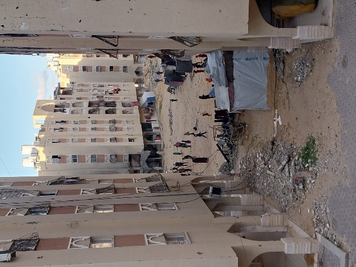 The image depicts an elevated view of a densely populated, damaged residential area in Khan Yunis, Palestinian Territory. Multiple multi-story apartment buildings, constructed primarily with light beige facades, enclose an open, unpaved communal space. These buildings exhibit extensive external damage, including numerous impact marks consistent with bullet holes or shrapnel on their walls, and many windows are broken, shattered, or missing panes, with some covered by makeshift materials or twisted metal security grates. Clothes are visibly hanging from numerous balconies and windows across the structures.
The ground between the buildings consists largely of disturbed earth and sand, littered with significant amounts of rubble, concrete fragments, and assorted debris. In the central foreground, a large pile of charred, twisted metal and other destroyed materials is prominent, suggesting a recent localized impact or fire. To the right of this debris, a makeshift shelter constructed from light grey tarpaulin, bearing visible UNICEF logos, stands on the dirt ground, with additional bedding or clothing draped over its roof. A smaller, darker structure is partially visible behind it, and another blue tent-like structure is further in the background.
Approximately 20-30 individuals, including adults and children, are scattered across the open dirt area, moving or standing in small groups or individually. They appear to be civilians, dressed in various modest clothing. No specific interactions or detailed actions are clearly discernible beyond general movement through the area. Exposed electrical wires or cables hang from the upper levels of the building on the left. The sky above is clear with scattered clouds.