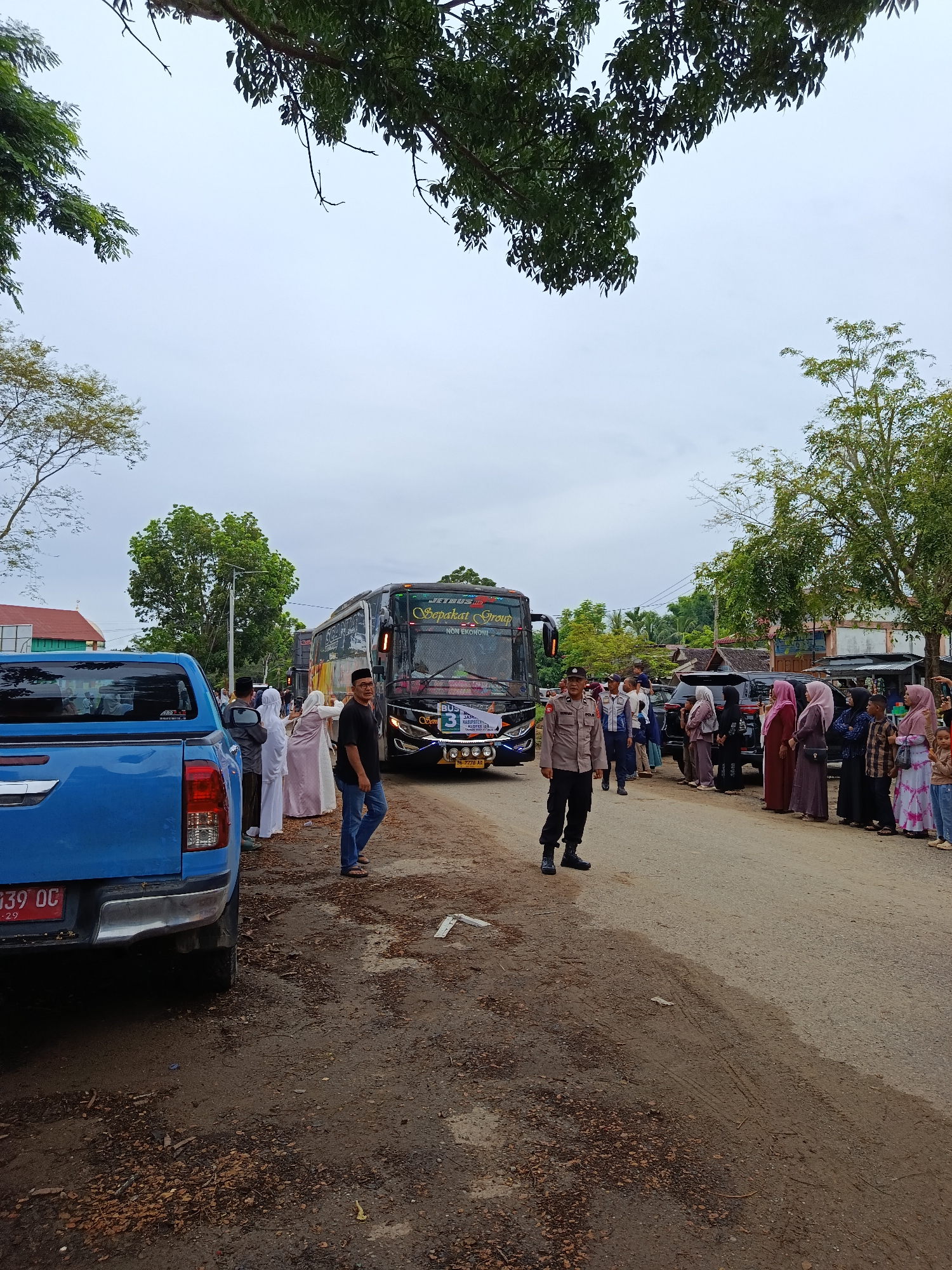 A group of people are gathered on a dirt road in Meureudu, Indonesia, near a large bus. The bus is black with colorful decals and reads "JETBUS" and "Sepsaka" above the windshield. A blue pickup truck is parked on the left side of the road, its rear visible. Several people are standing in a line on the right side of the road, some wearing traditional Islamic clothing. A police officer in uniform is standing near the bus, looking towards the camera. The sky is overcast, suggesting a cloudy or possibly rainy day. The trees are lush and green, indicating a tropical or subtropical climate. The ground is a mix of dirt, gravel, and scattered dry leaves. The scene appears to be a pickup or drop-off point for the bus, possibly a group traveling together. The presence of the police officer suggests an organized event or a need for security. The mood appears calm and orderly. There is text on the license plate of the truck, "B 939 QC", and on the bus, including "JETBUS", "Sepsaka", and a number "3". There is also text on a banner on the front of the bus, though it is difficult to read.