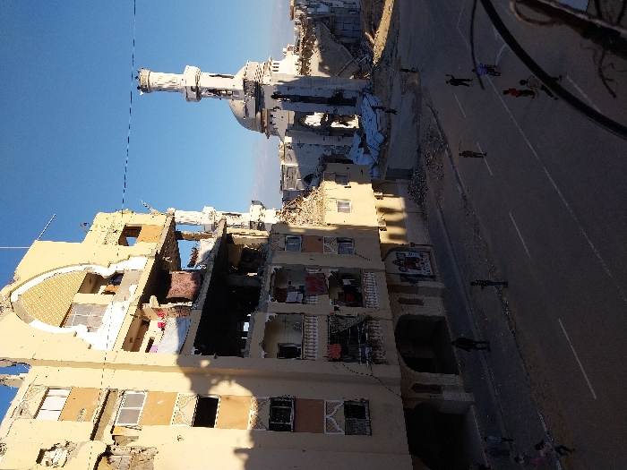 This image depicts a devastated urban street scene in Khan Yunis, Palestinian Territory. The foreground features a multi-story building on the left, severely damaged with large sections of its facade and interior exposed. Multiple floors show blown-out walls, shattered windows, and visible debris. Clothes are seen hanging on a lower balcony. To the right and behind this building stands a mosque, also exhibiting significant structural damage, particularly to its lower sections, though its white dome and minaret remain largely intact against a clear blue sky.
The paved asphalt street in the mid-ground has white lane markings. Several individuals, primarily children, are scattered across the street and sidewalk. A child on a bicycle is visible, and other children appear to be walking or playing. A few adult figures are standing on the street and sidewalk. Debris piles are visible on the ground near the damaged structures. Long shadows are cast from the buildings onto the street, suggesting either morning or late afternoon. A thin black wire crosses horizontally in the upper middle of the frame. The photograph appears to be taken from an elevated position, indicated by a partial railing visible in the bottom right corner.