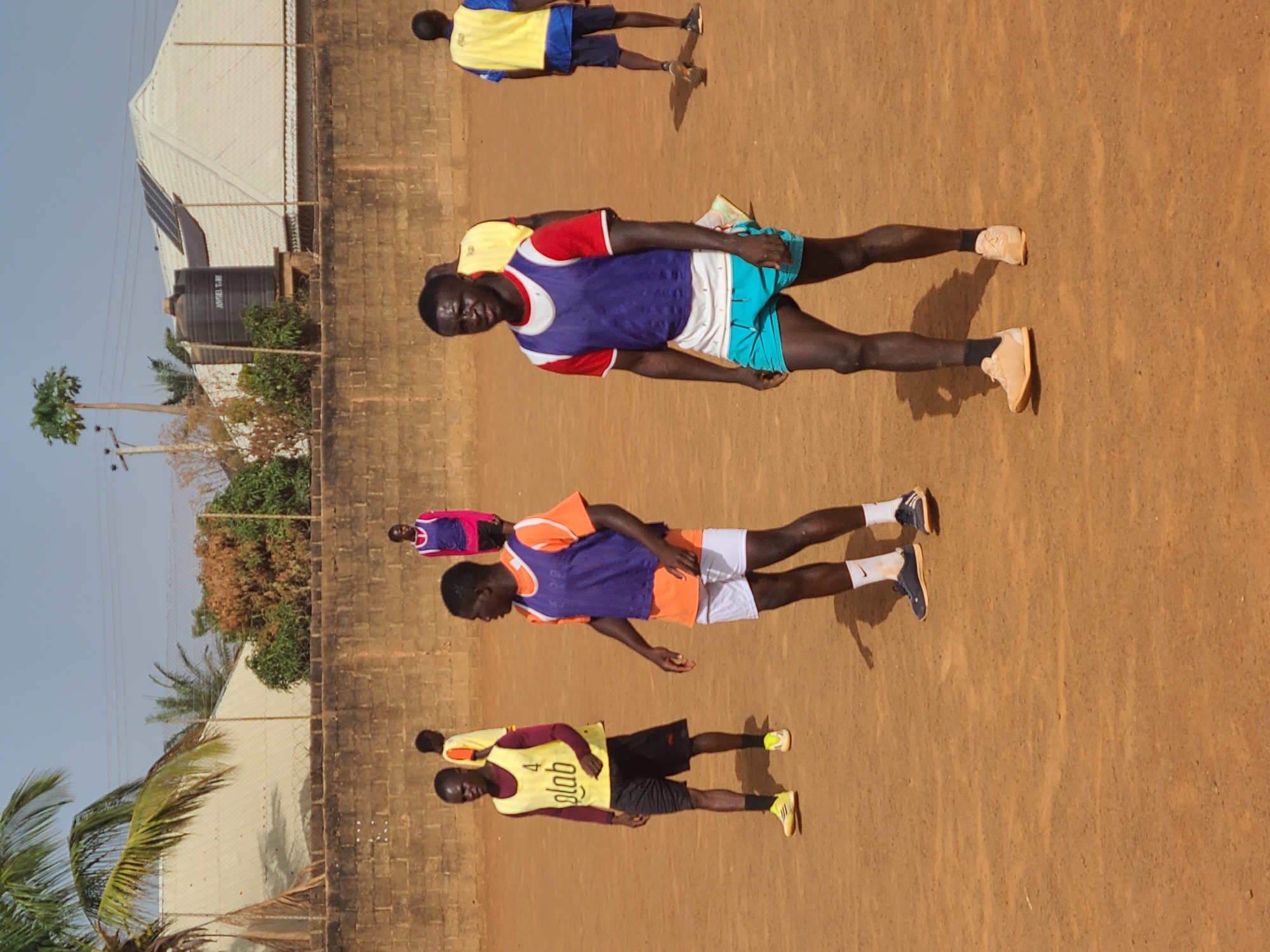 Captured at Barakin Lerawi, Nigeria, the image depicts a group of young men on a reddish-brown dirt field. They appear to be involved in some form of athletic activity, possibly a practice or training session. The individuals are wearing various athletic jerseys and shorts, with some in yellow and others in combinations of blue, orange, red, white, and purple. A low brick wall runs along one side of the field. In the background, there is a building with a light-colored roof and a dark water tank. A few trees, including a palm tree, are visible. The sky is clear and blue.