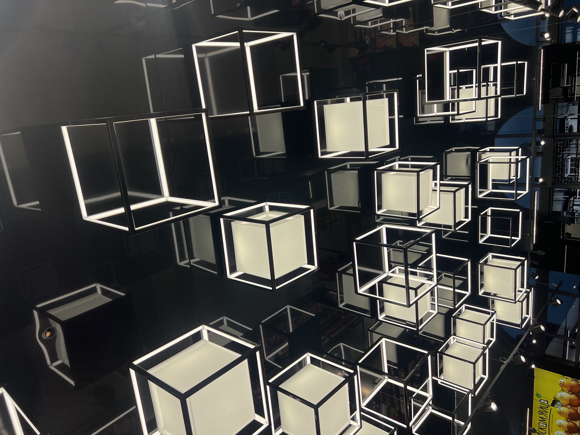 The image shows a dense arrangement of illuminated, cubic light fixtures suspended from a highly reflective, dark ceiling. The cubes vary in design; some consist only of illuminated edges, while others have opaque panels on their faces, giving off a diffuse light. The reflections on the ceiling create a disorienting effect, making it hard to discern the actual spatial arrangement. The setting seems to be inside an interior space, possibly a commercial or exhibition area. The exact location is unknown.
