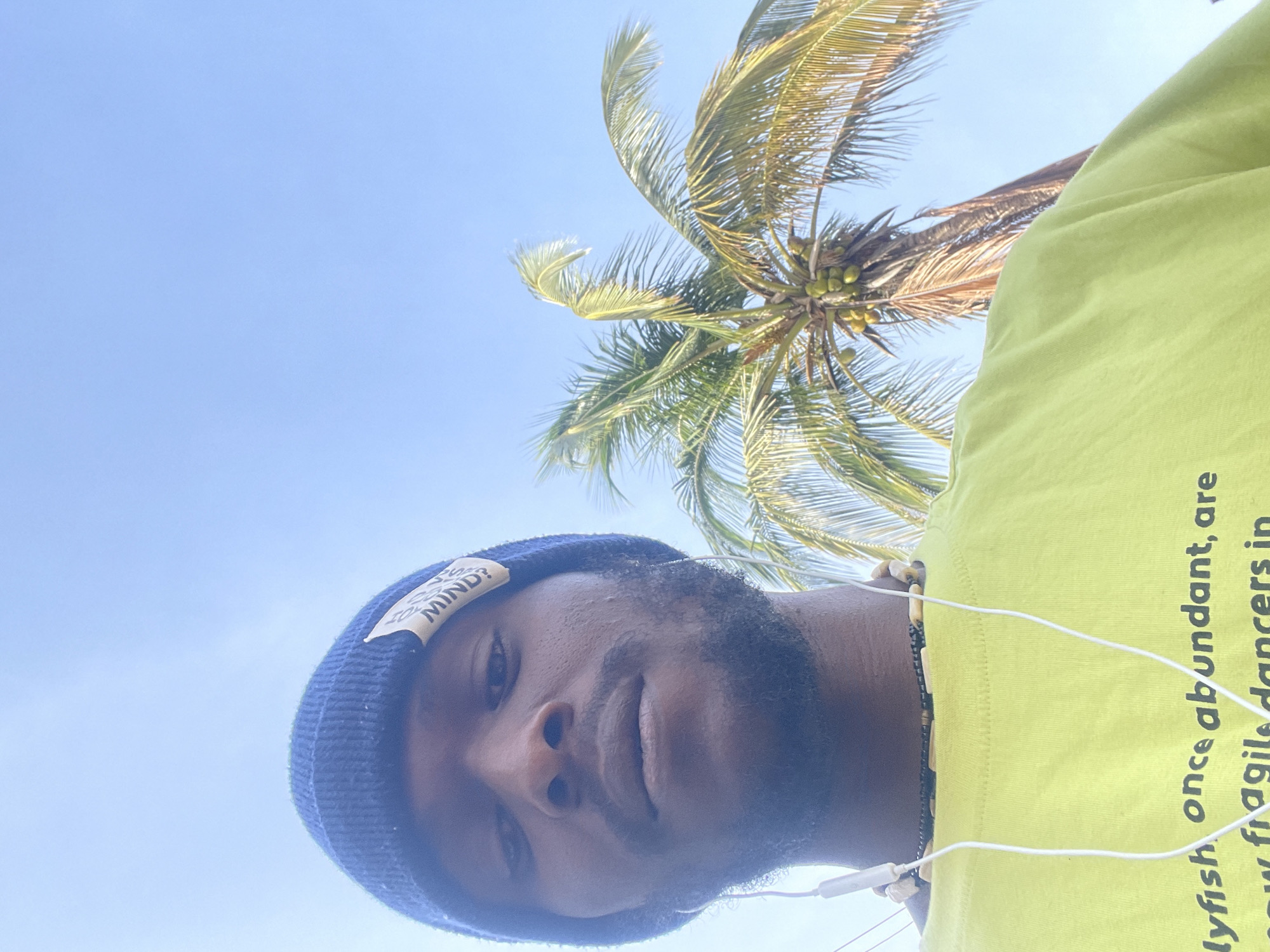 A low-angle shot captures a Black male wearing a dark blue beanie, a light green shirt, and white earphones. A palm tree is behind and to the right. The sky is light blue. It is not possible to confirm the location is in Jos, Nigeria, based on visual information alone.