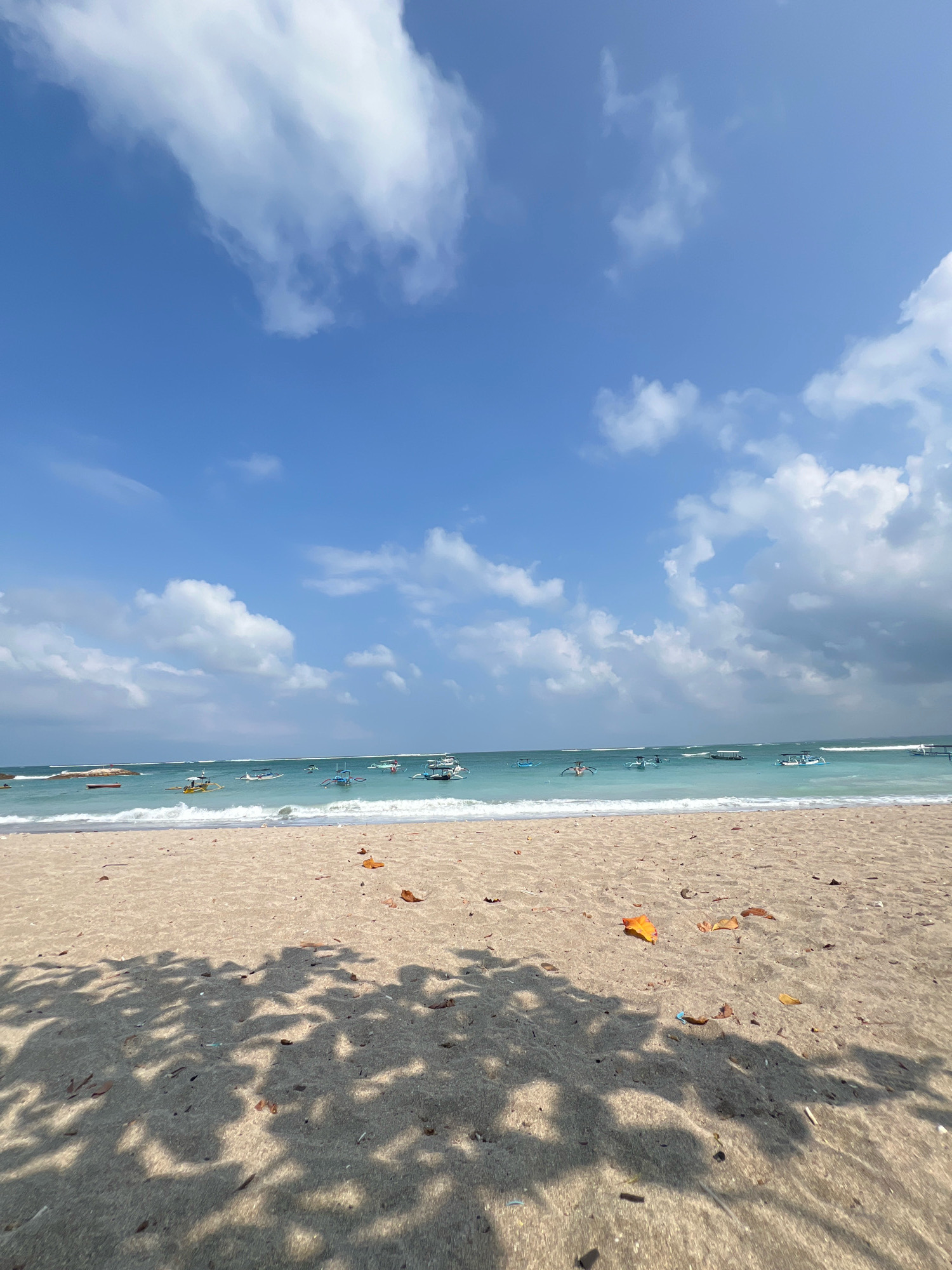 A wide-angle shot captures a serene beach scene in Kuta, Indonesia. The foreground is dominated by a sandy beach, with patches of dry leaves scattered across the sand, and shadows from overhead foliage creating a dappled pattern. The midground features the gentle turquoise waves of the ocean lapping onto the shore, and in the distance, numerous traditional Indonesian fishing boats are anchored on the calm water. The background is a vast expanse of blue sky, dotted with fluffy white clouds, suggesting a bright and pleasant day. The overall impression is one of tranquility and natural beauty.