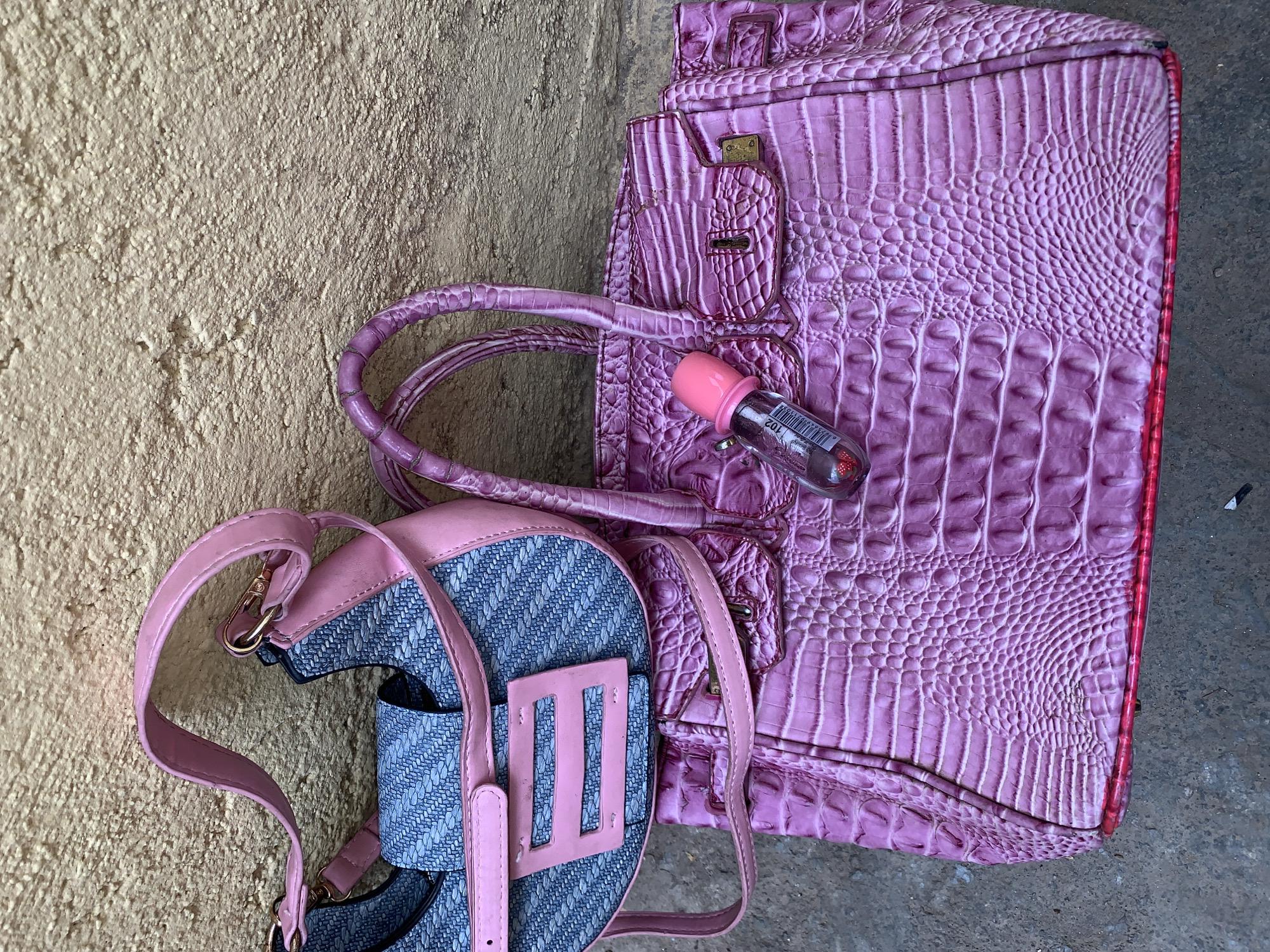 The image shows a pink alligator-skin-patterned handbag and a pink-strapped backpack lying on what appears to be a weathered, light-brown concrete or stone surface. On the handbag sits a small, clear bottle with a pink cap. The backpack is predominantly blue with a pink trim and buckle. The location is not identifiable from the image. No people, actions, or interactions are depicted.
