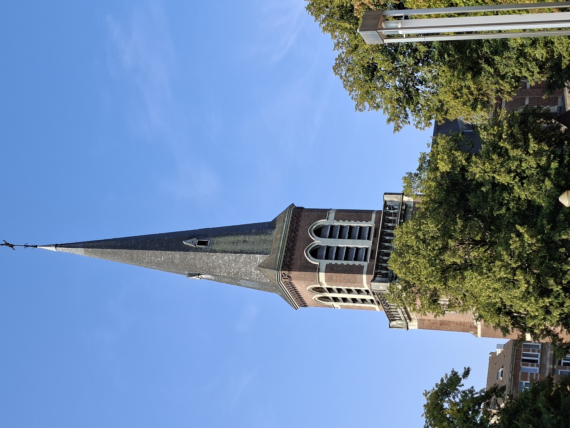 The image shows the spire of a brick church against a clear blue sky. The spire is tall and pointed, covered in grey tiles, and topped with a cross. The body of the church is made of red brick and features arched windows. Lush green trees partially obscure the view of the church's lower sections. To the left, the roof of another building is visible, and to the right, a modern street lamp with multiple vertical poles stands among the trees. The scene appears to be an outdoor urban environment, likely during daylight hours with good weather. The specific location in Brussels, Belgium, is not discernible from the image alone.