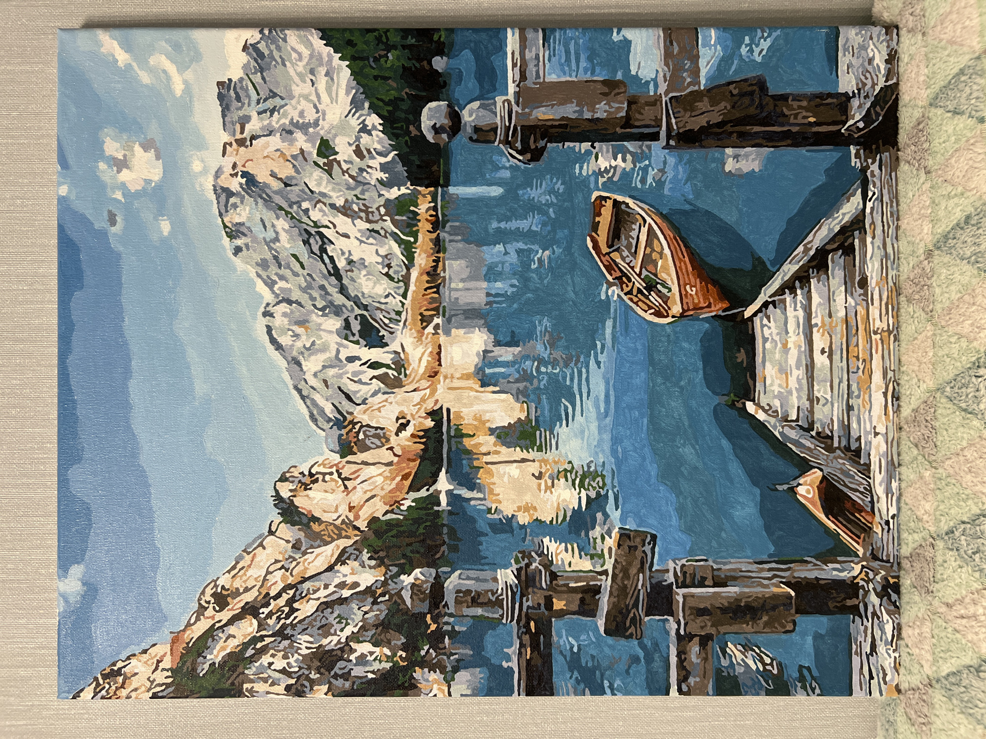 The image depicts a painting of a landscape featuring a mountain range, a lake, and wooden docks. A rowboat is visible on the water near the docks. The scene is likely inspired by a natural environment, possibly a mountain lake setting. The style is representational with a somewhat simplified, painterly technique. The location in the city of Vnukovo, Russia, is not represented.