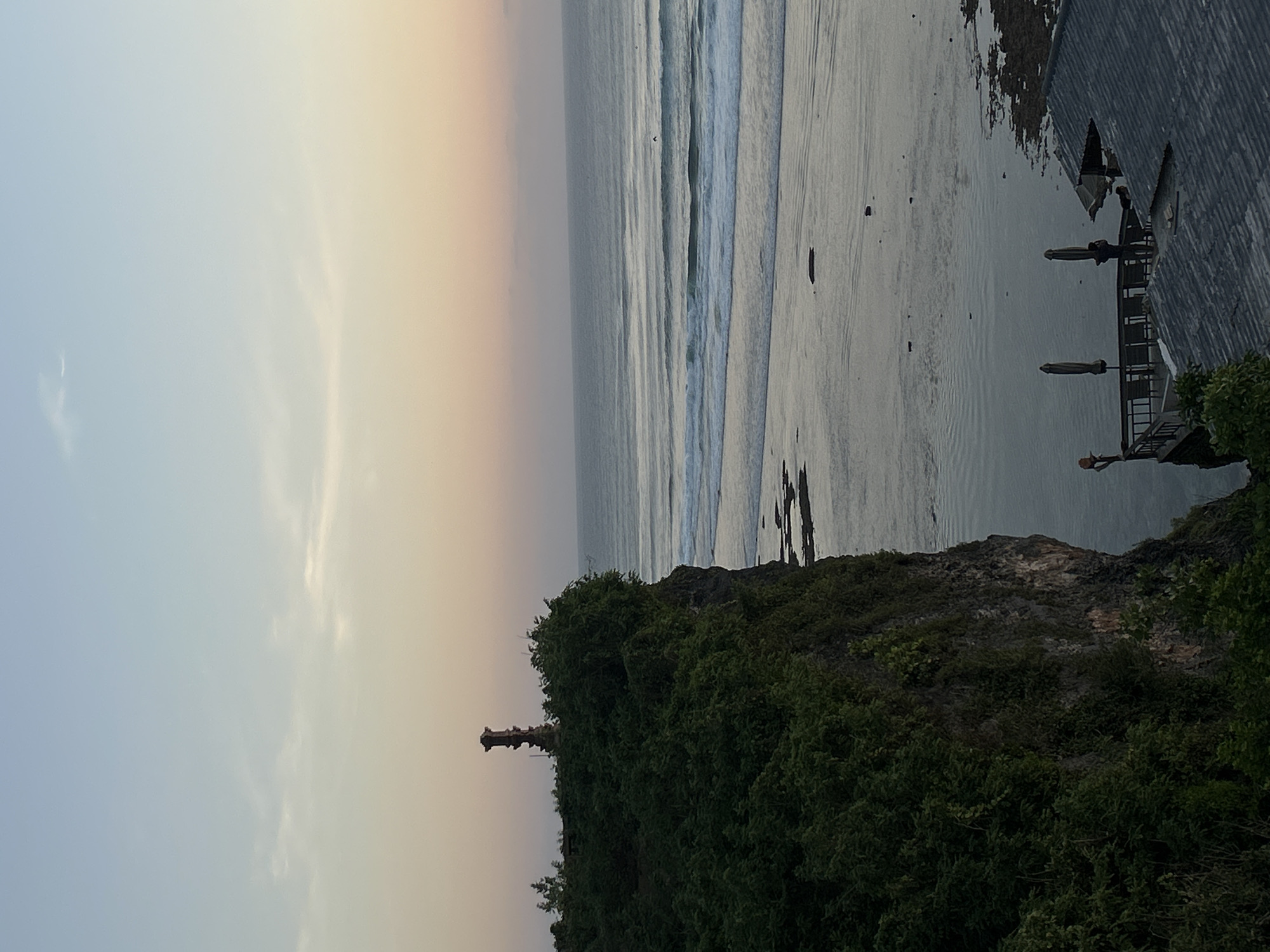 The image depicts a scenic view of a cliff overlooking the ocean at sunset in Kuta Selatan, Indonesia. A dark, lush green cliff face dominates the left side of the frame, topped with dense foliage and a weathered stone monument. The ocean stretches to the horizon, with gentle waves breaking on a sandy beach. In the foreground, a rooftop with dark tiles and a balcony area with tables and umbrellas suggests a dining or viewing establishment. A lone figure stands on the balcony, seemingly observing the sunset. The sky transitions from a pale blue at the top to a soft orange and pink hue near the horizon, indicating the approach of evening.