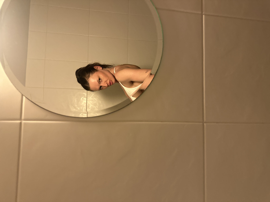 A person, reflected in a round, slightly beveled mirror, appears to be peering around the edge. She has dark hair pulled back and wears a light-colored top. The mirror is mounted on a tiled wall. The tiles are square and a neutral, light brown color with darker grout lines. The lighting in the room is warm, casting a subtle glow on the tiles. The arrangement gives the impression of a candid or intimate moment captured.