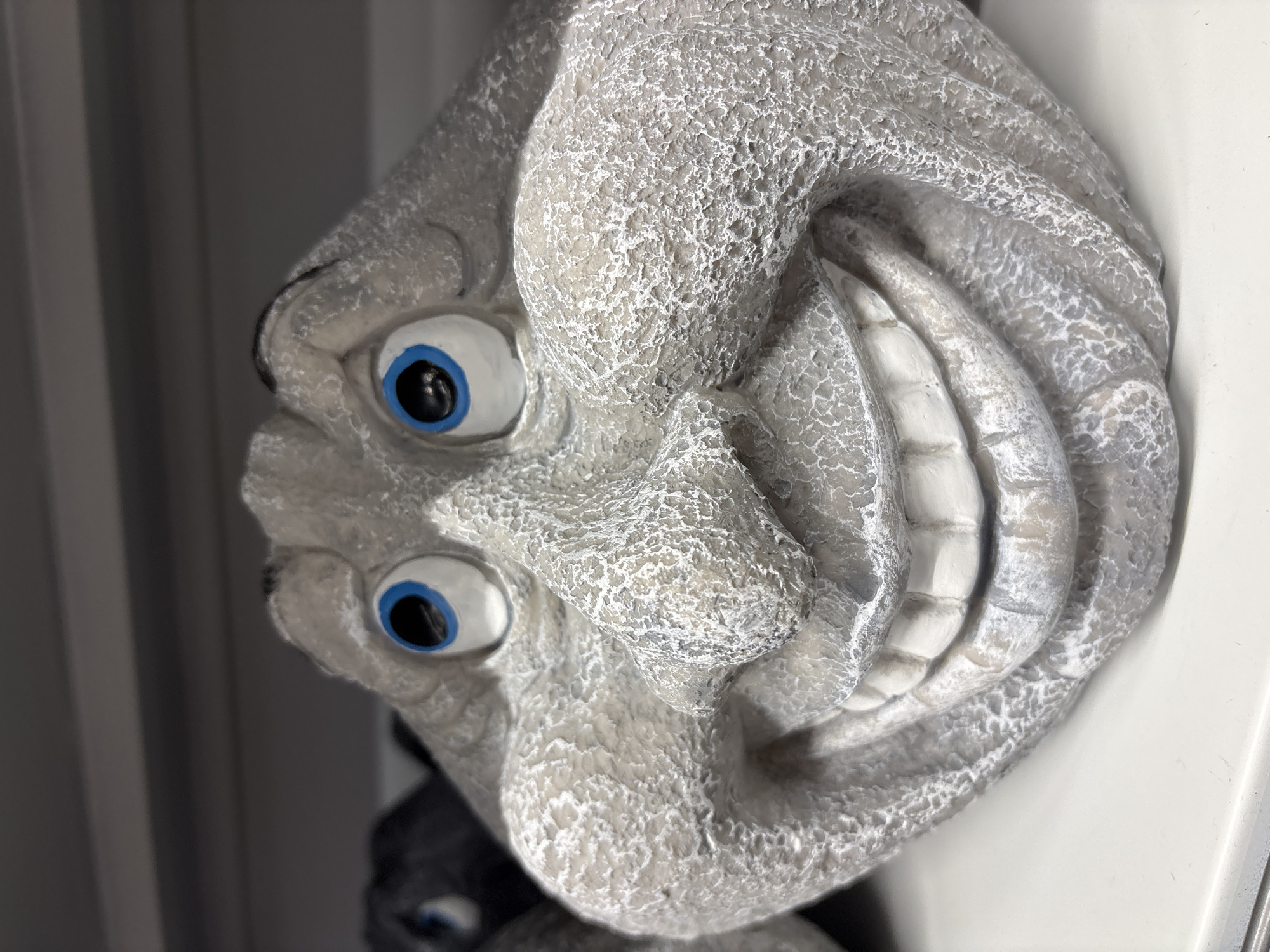 A close-up shot features a whimsical stone garden statue with a large, smiling face. The statue, primarily grey with white textured details, has prominent blue eyes with black pupils, a broad nose, and an exaggerated grin showing rows of white teeth. The texture of the stone suggests it is weathered or designed to look that way, with a rough, pitted surface. In the background, another dark-colored statue is partially visible, also with blue eyes. The statues are placed on a white shelf or surface, with horizontal grey shelving above. The overall lighting is neutral, highlighting the statue's features without harsh shadows. The image appears to be taken indoors, possibly in a store or a display area, and there is no indication of a specific location within the city of València, Spain.