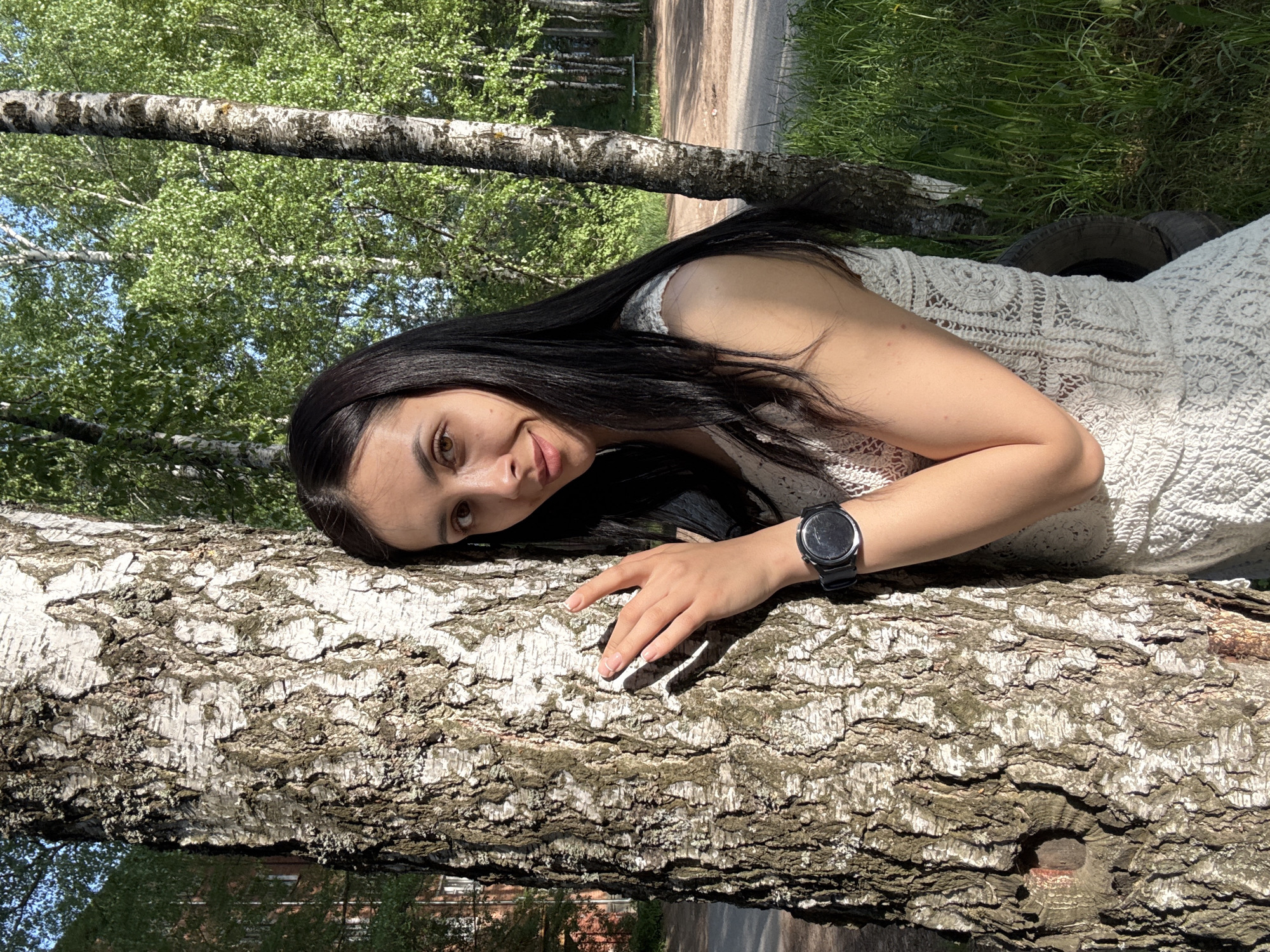A young woman lies on a fallen birch tree. She is wearing a white dress and a black smartwatch. She is resting her head and hand on the tree trunk, gazing towards the camera with a slight smile. The location is Votkinsk, Russia, as suggested by the presence of birch trees, commonly found in the region. The background features green grass, other trees, and a path.