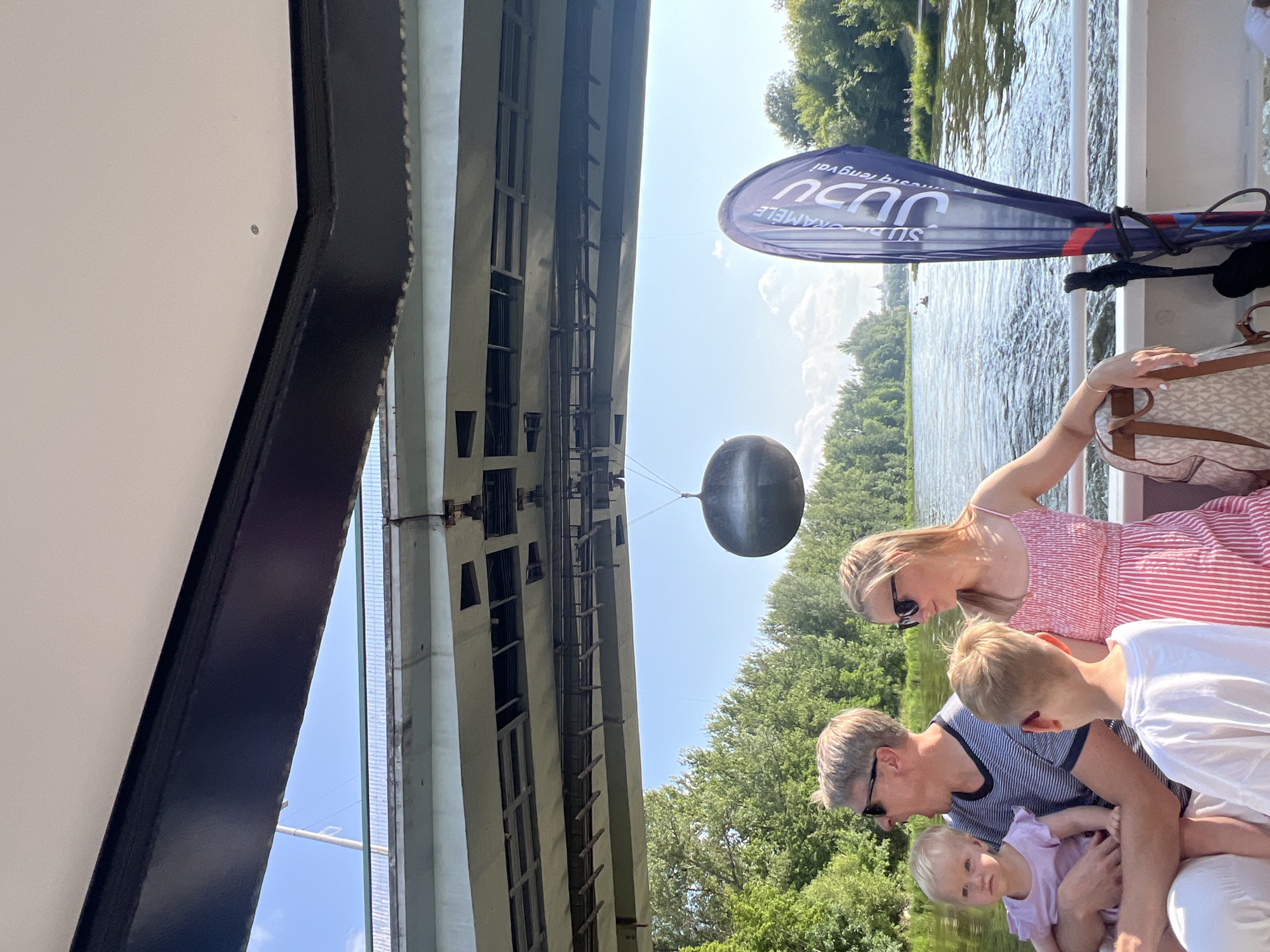 In Vilnius, Lithuania, a family of four is enjoying a boat ride on the Neris River, passing under a large bridge. The father, wearing sunglasses and a striped t-shirt, holds a young child in a light purple dress. Their son, wearing a white t-shirt, stands beside them. The mother, in a pink striped dress and sunglasses, stands next to the father. A dark, spherical object hangs suspended in the air from a thin wire beneath the bridge. A blue flag with white text, partially obscured, stands upright on the boat. The river is calm, with lush green trees lining the banks. The sky is clear and blue with a few scattered clouds.
