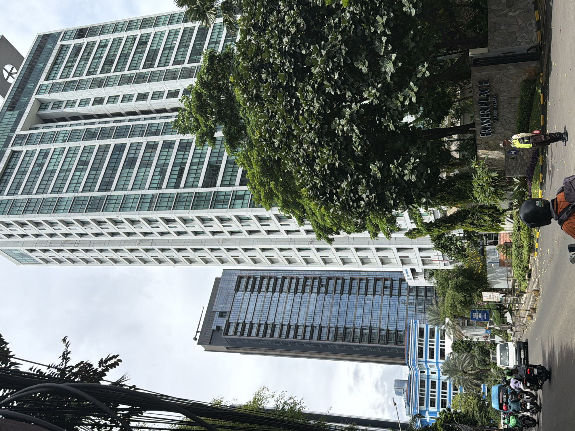 The image depicts an urban landscape, specifically Fraser Place Setiabudi Jakarta, located in South Jakarta, Indonesia. A tall, white building with numerous windows dominates the left side of the frame. To the right of this building, a large, full tree partially obscures a sign indicating "Fraser Place." A uniformed individual is standing near the sign. In the lower part of the frame, a darker, glass-fronted building is visible. The street is occupied by several motorcycles and a white vehicle. Overhanging wires and trees are visible at the bottom. The sky is overcast.
