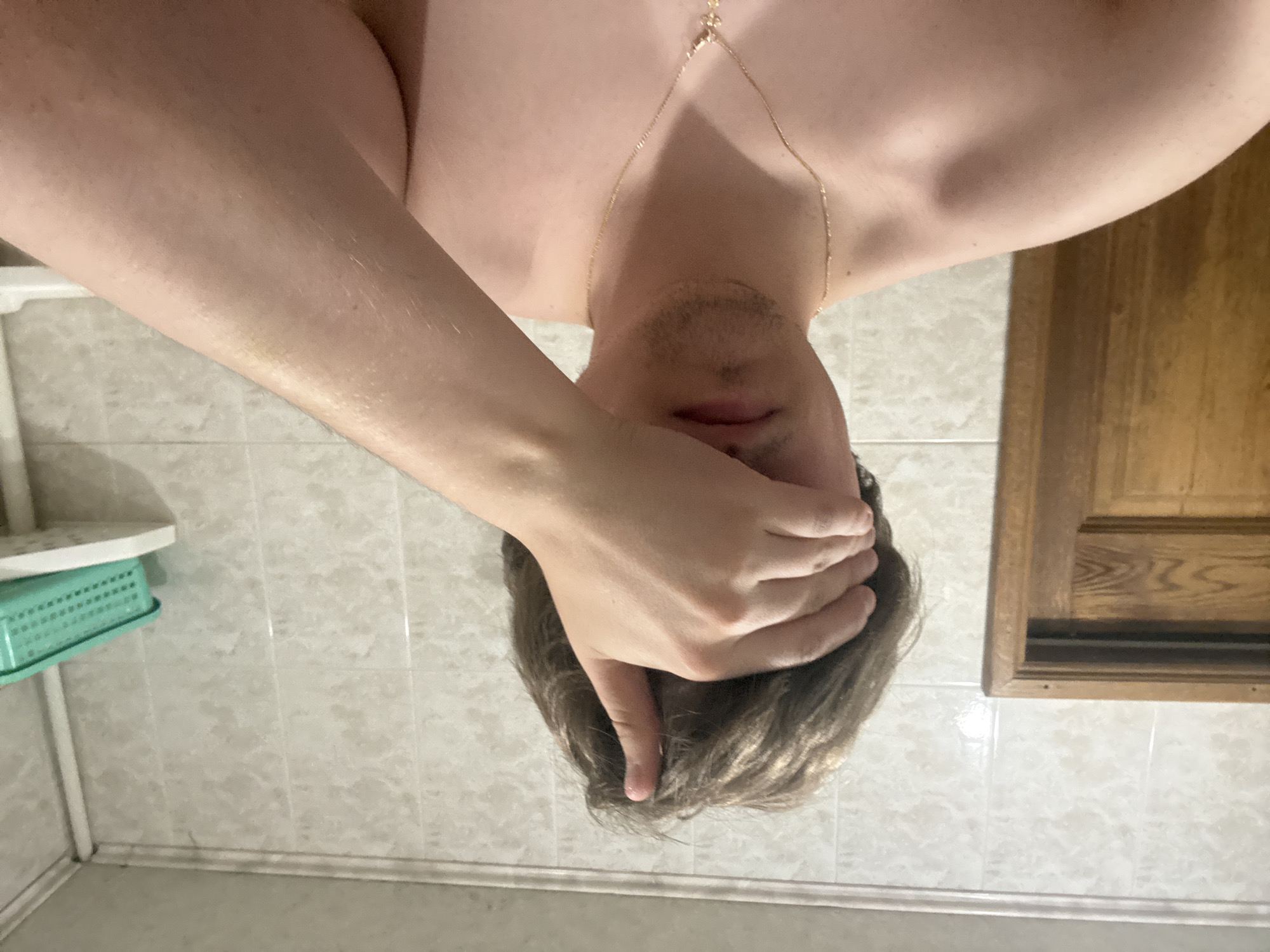 A young man is taking a selfie in a bathroom. He has his right hand covering his eyes. He is wearing a gold chain with a cross pendant. The bathroom has tiled walls and a wooden door. A green plastic basket is visible on a shelf in the background. The photo is taken from the chest up.