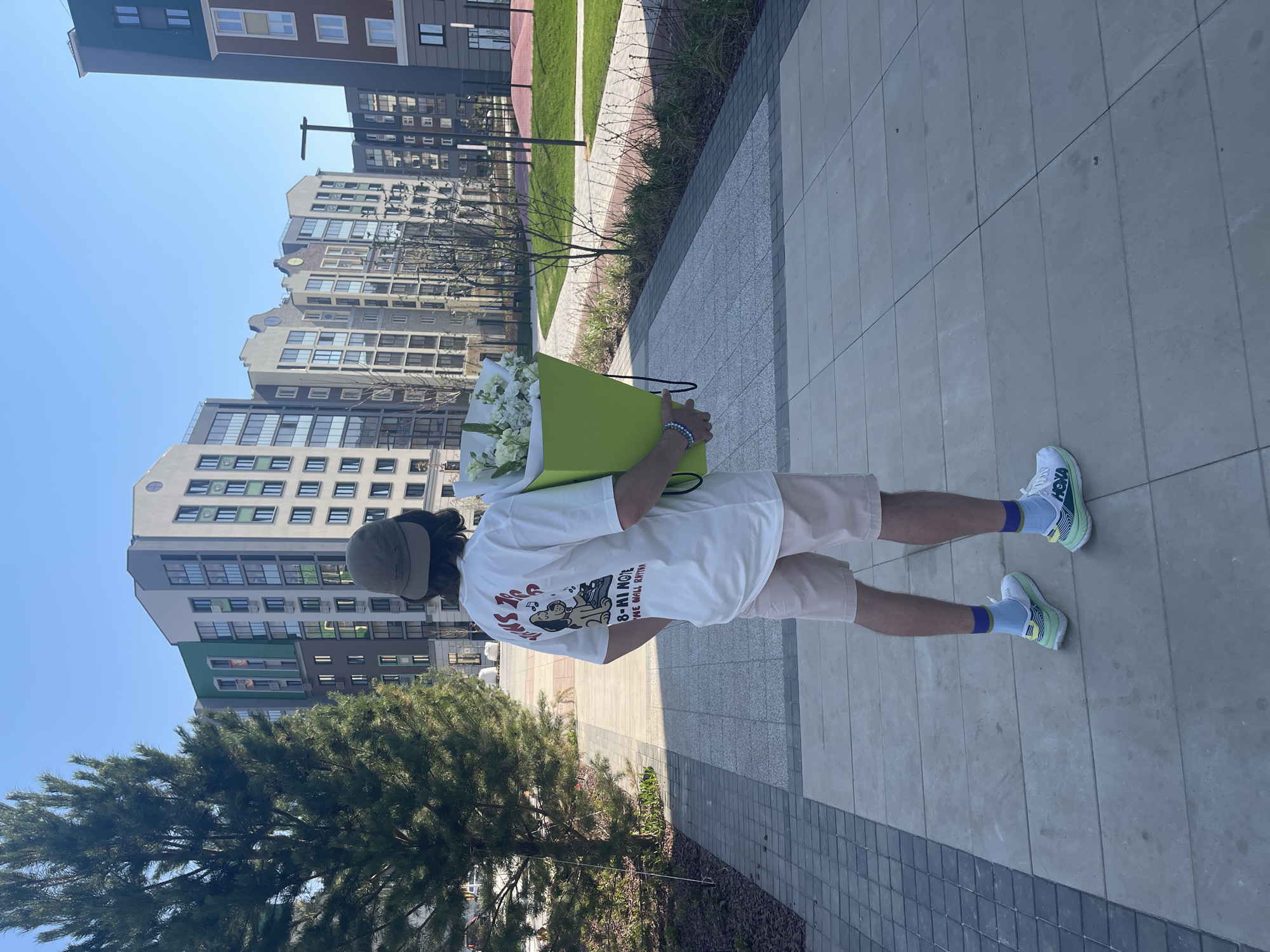 A person is carrying a large bouquet of white flowers in a green bag while walking on a paved sidewalk in what appears to be an urban area. The person is wearing a white t-shirt with a graphic on the back, light pink shorts, light blue socks with blue and gold details, and white running shoes with green accents. They are also wearing a hat that obscures their face. The background features modern apartment buildings with a mix of gray, beige, and green facades. A young evergreen tree and some landscaping are visible on the left. The image suggests a sunny day with a clear blue sky. The location may be Хохряки, Russia, as specified.