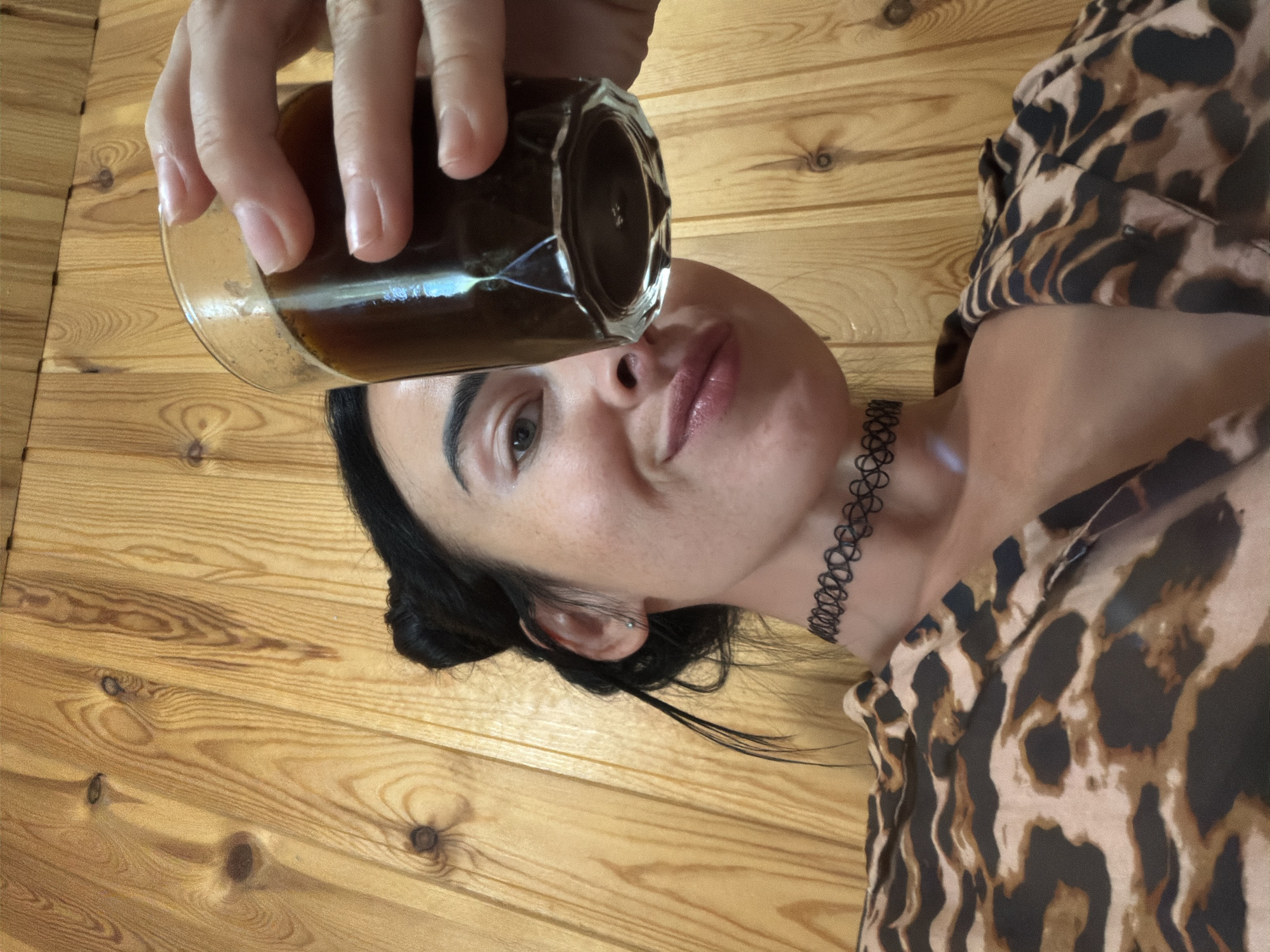 The image shows a close-up, low-angle shot of a woman holding a glass of dark liquid, presumably coffee, up to the camera. She has dark hair pulled up in two buns, visible strands of hair framing her face. She is wearing a leopard-print top and a black choker necklace. Her facial expression is a slight smile, and she is looking towards the camera. The background consists of a wooden wall with vertical planks, suggestive of an interior setting like a cabin or a sauna. There are no identifiable landmarks or features that would indicate the specific location within Sochi, Russia. The lighting is moderate, highlighting the woman and the drink, with some reflection on the glass.