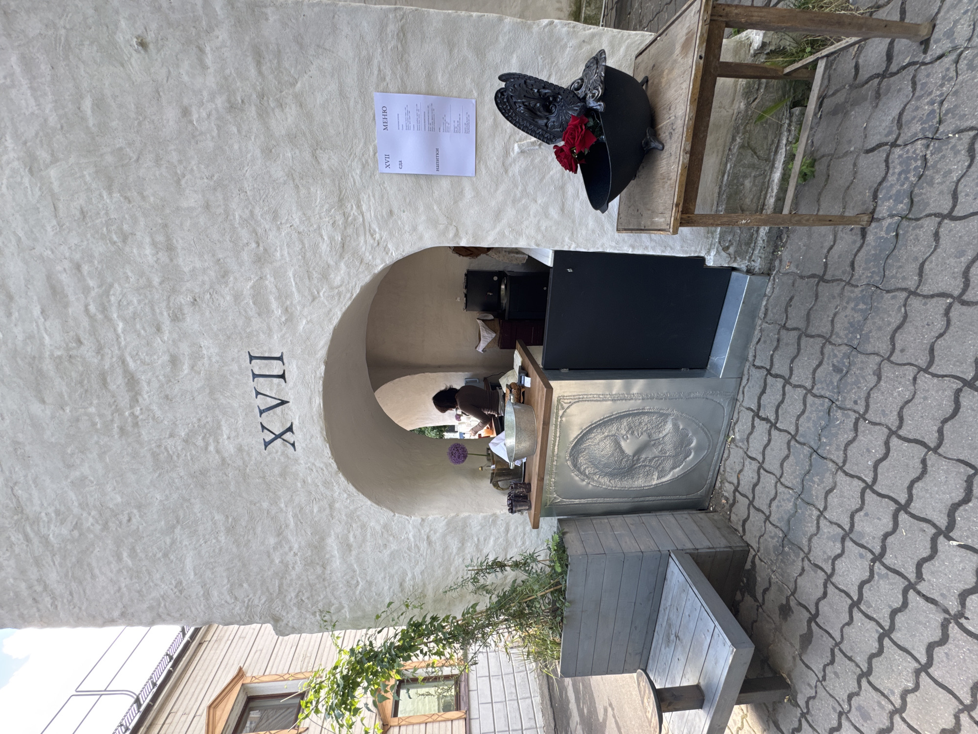 The image depicts the exterior of a small establishment with a white, stucco-textured wall featuring a large arched opening. Above the arch, the Roman numeral "XVII" is prominently displayed. To the right of the arch, a white paper menu is affixed to the wall. Inside the arch, a person is visible behind a counter, appearing to be working. The counter itself is metallic with an embossed circular design. In front of the establishment, to the left, is a long, grey wooden planter box with a green vine growing out of it, and a small wooden bench with a circular table top. To the right of the arch, a small, rustic wooden table holds a black decorative object resembling a small bathtub filled with red flowers. The ground is paved with interlocking grey tiles. The architectural style and the signage suggest this could be a cafe or a similar establishment in an urban setting. The presence of the menu in Russian indicates a location in Russia. The image was taken in Moscow, Russia.