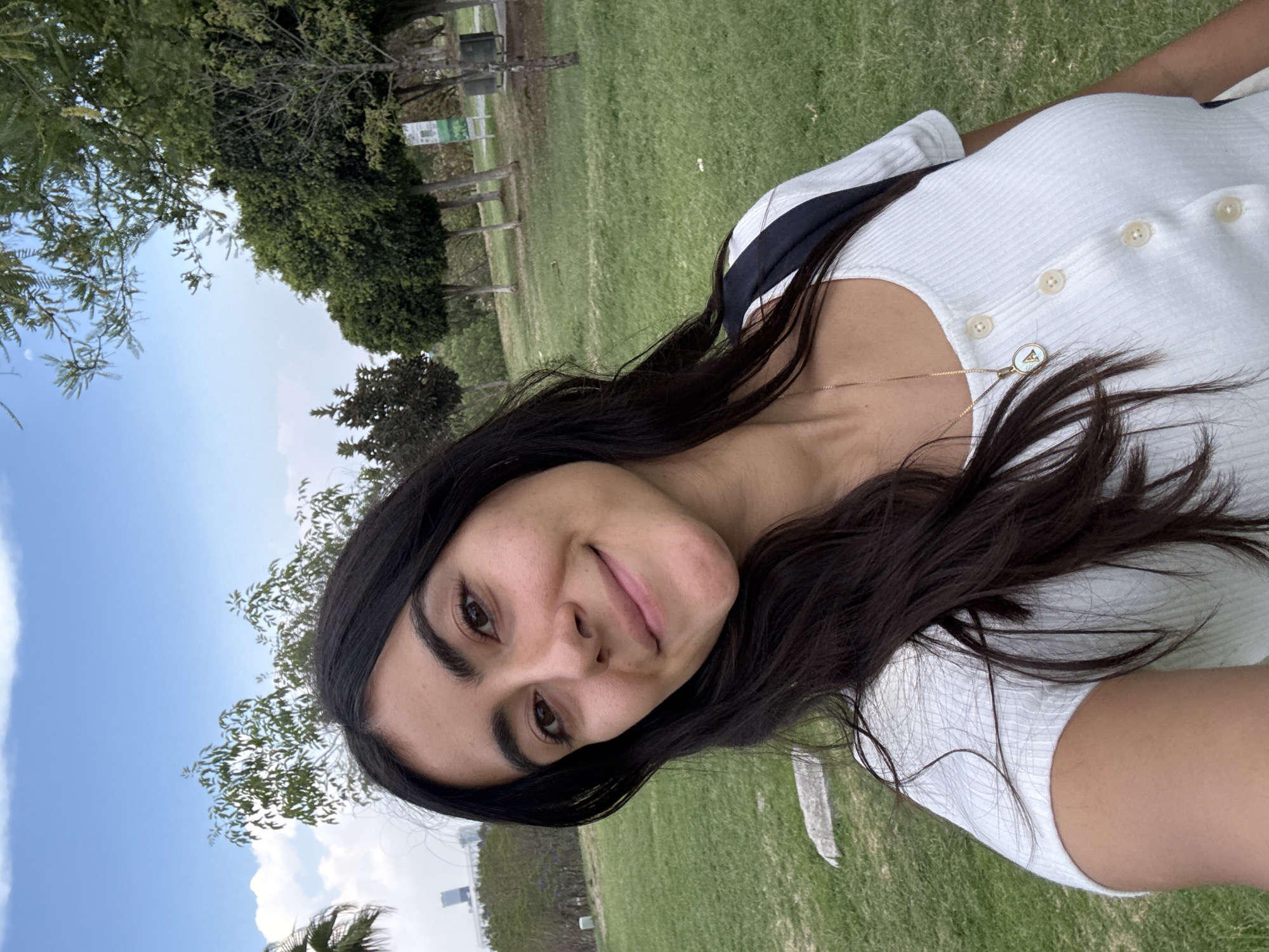 A young woman with dark hair, wearing a white top with buttons, is positioned at an angle in the foreground of the image. She has a necklace on. The background consists of a grassy area, trees, and a blue sky with clouds. In the distance, what appears to be a building can be observed. Given the location of Puebla, Mexico, it is plausible she is in a city park, or a field.
