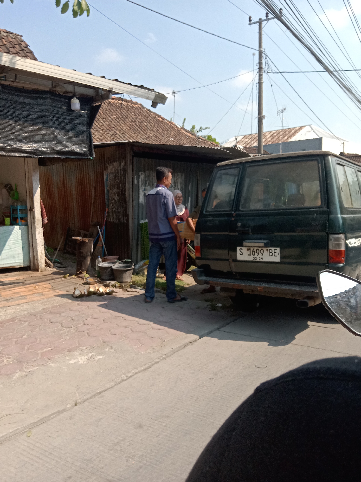 In Sugihwaras, Indonesia, on a sunny day, a dark green van is parked on the right side of the image. The van's license plate reads "S 1699 BE". A man in a blue shirt and jeans stands to the left of the van, facing away from the camera. A woman in a brown headscarf and long brown dress stands next to him, facing towards him. In front of them, on a cobblestone path, are several halved coconuts and two buckets. To the left of the man is a building with a corrugated metal wall and a dark tarp overhead. A street lamp is visible on a pole in the background, with numerous electrical wires crisscrossing the sky. The ground is paved with a mix of cobblestones and concrete. The image is taken from a low angle, possibly from a motorcycle, as evidenced by the reflection of a mirror in the lower right corner.