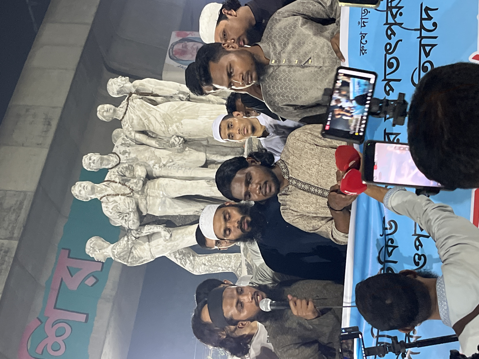 A group of men are gathered in front of the Aparajeyo Bangla sculpture at the University of Dhaka in Dhaka, Bangladesh. One man is speaking into a microphone. Several others are filming the event with smartphones. A banner with text in Bengali is visible.