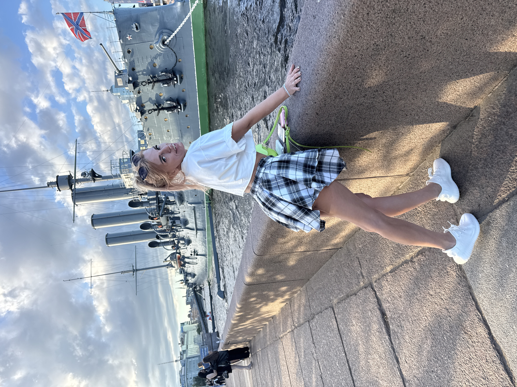 A young Caucasian woman with blonde hair pulled back, wearing a white cropped t-shirt and a black and white plaid skirt, poses on a stone embankment. She has sunglasses perched on her head and is leaning on the embankment wall. Behind her, a large gray warship, identified as the Aurora cruiser, is docked in the Neva River. The ship features multiple smokestacks, masts, and cannons, with a Russian flag flying from its stern. In the background, city buildings line the riverbank under a cloudy sky. Two other women in dark clothing are visible further down the embankment, walking away from the camera. The lighting suggests it is late afternoon or early evening.