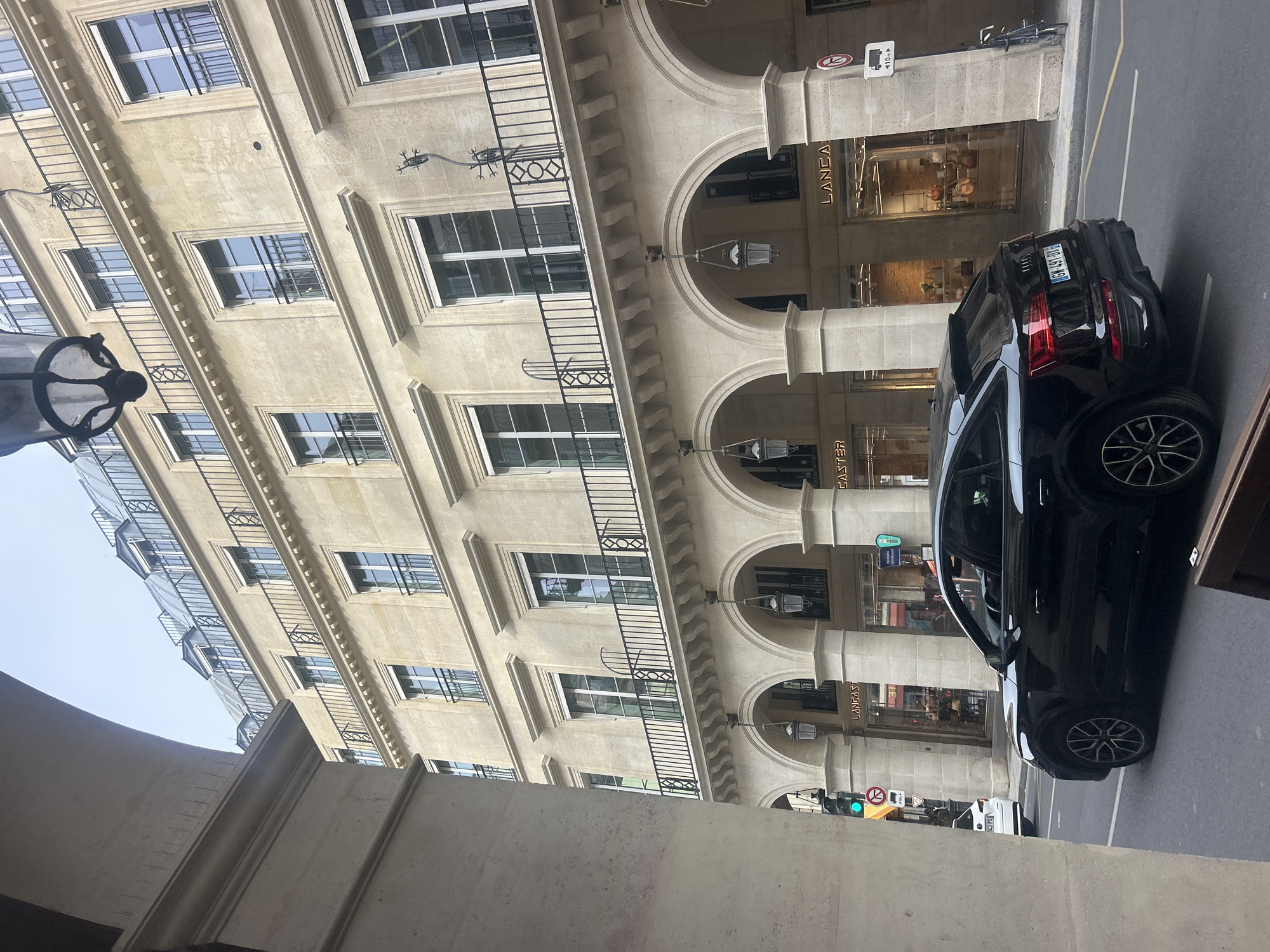 Here is a description of the image:
The image depicts a street scene with a light-colored building featuring arched colonnades and black iron railings in Unknown, Unknown. A black car is parked on the right side of the frame, facing towards the foreground. The car has a European license plate. The building houses shops, including one identified by signage as "LANCASTER." Streetlights are visible, and traffic lights indicate a green signal. The perspective of the photograph is partially obstructed by a concrete structure in the lower left corner, which seems to be a part of an archway or colonnade. The weather appears overcast.