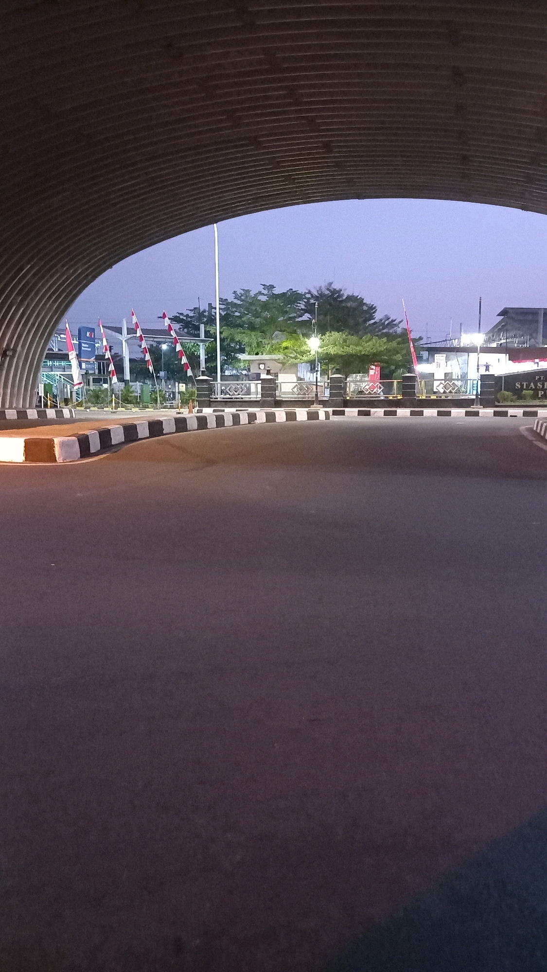 The image shows a view from under a large, curved, ribbed metal structure that appears to be a tunnel or underpass. The foreground is dominated by a paved road with a slight curve to the right. The road surface is dark asphalt with white and black painted markings along the edge of a curb. Beyond the road, a barrier with black and white striped bollards separates the road from a pedestrian area.
In the middle ground, several Indonesian flags (red and white) are visible, mounted on poles. Behind the flags, there are trees and some buildings. One building appears to have signage that reads "PT. STASI..." which likely indicates "PT. Stasiun" or "PT. Station," suggesting a train station. There are also streetlights and some other structures that resemble buildings or infrastructure. The sky is a pale, dusky blue, indicating it is either early morning or late evening.
The scene appears to be located in the city of Surakarta, Indonesia, specifically near a train station as suggested by the signage. There are no people visible in the image. The overall impression is one of an urban environment at dusk or dawn, with the infrastructure of transportation being a prominent feature.