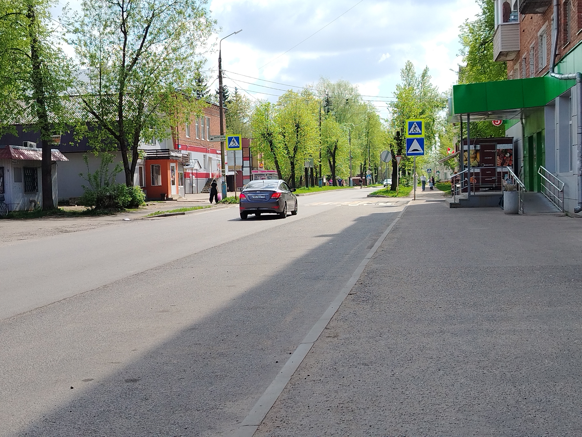 The image depicts a street scene in an urban area. On the left side of the street, there is a gray building with a red roof extension, followed by a small orange structure. Further along, a brick building is visible. On the right side of the street, a building with a green awning stands. The street itself is gray and slightly cracked, with a curb separating it from the sidewalk.
A gray car is driving on the street away from the viewer. A person is walking on the left side of the street, near the orange structure. A pedestrian crossing sign is visible on both sides of the street. Trees with green leaves line the street. The sky is partly cloudy.
