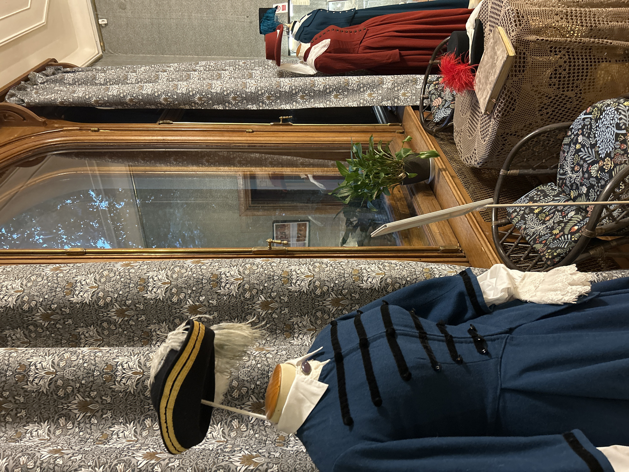 The image depicts a display of historical costumes in what appears to be a museum or historical house exhibit. In the foreground, a mannequin is dressed in a blue military-style coat with black trim and braiding, a white ruffled collar, and lace cuffs. It wears a black bicorne hat adorned with gold trim and white feathers. Behind this mannequin, and further to the right, are two more mannequins. The one on the left wears a long, red historical dress with a ruffled collar and a red hat. The mannequin on the right is dressed in a teal-colored historical dress with a white collar.
The mannequins are positioned near a large, arched wooden-framed window. Through the window, a reflection of trees and sky is visible. A potted plant sits on the windowsill. In front of the window and to the right of the mannequins are two wicker chairs with floral patterned cushions. A table covered with a crocheted lace tablecloth is situated between the chairs. On the table rests a closed book and a black hat topped with a vibrant red feather plume. The background features patterned wallpaper with a floral motif. The overall scene suggests a carefully curated exhibit designed to evoke a historical period. The location is not specified.