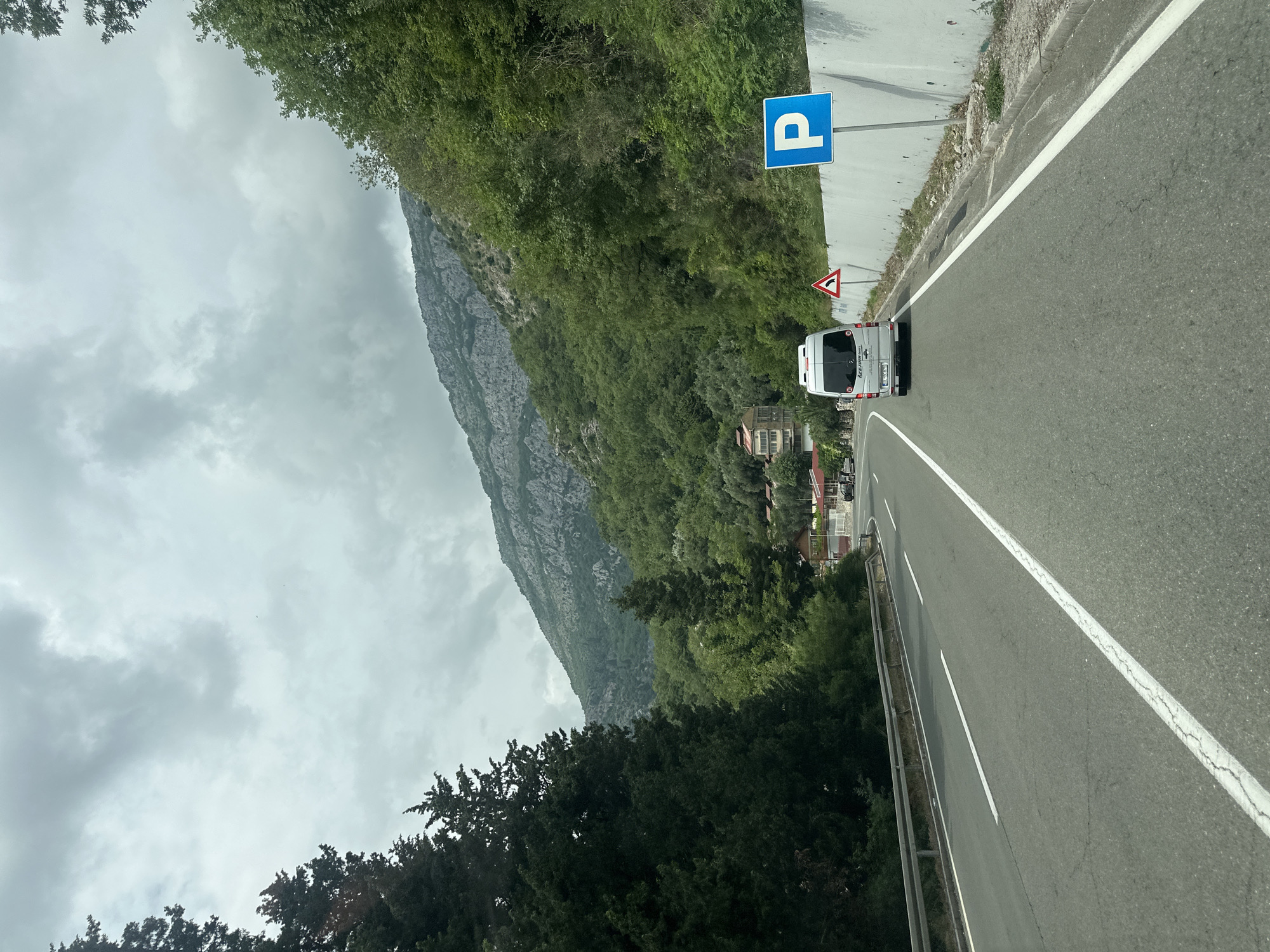 A white van drives on a paved road through a mountainous and heavily vegetated landscape under an overcast sky. The road, marked with white lines for lanes and a shoulder, curves gently. A metal guardrail runs along the left side of the road. On the right, a bright blue square sign with a white 'P' symbol indicates parking, positioned above a red triangular warning sign depicting a sharp left curve. Dense green trees and shrubs cover the hillsides along the road, partially obscuring residential buildings. A large, rocky mountain, also covered in patches of greenery, dominates the background on the left side of the frame. The sky is uniformly grey with thick clouds, suggesting an overcast day. On the rear of the white van, part of a brand name "LEPIS" is visible, along with letters "PG" on its license plate. The image captures a journey through a scenic, natural setting in Golubovići, Montenegro.