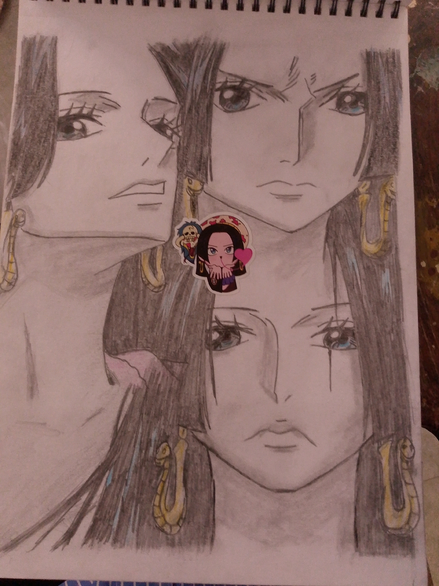 The image is a close-up drawing of three anime-style faces, layered and overlapping. The drawing is done on a spiral-bound notebook page. The faces have dark hair and prominent earrings shaped like snakes. A small sticker of a chibi character is placed on the upper right portion of the middle face. The drawing is primarily in grayscale with some colored accents on the sticker and earrings. The background of the image is the surface of a dark, textured table. There is no indication of the city of Sitarampuram, India, in the image or its contents.