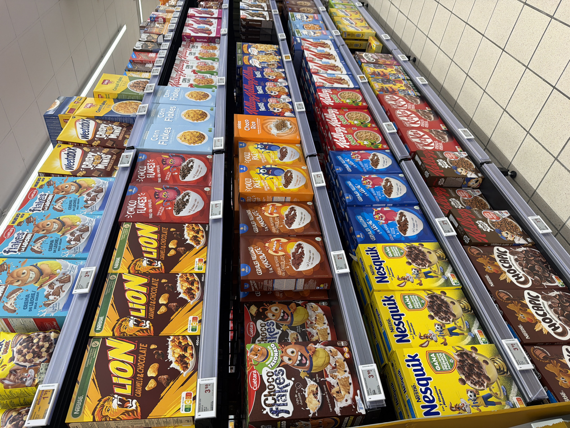 The image depicts a retail aisle in a supermarket, likely in the cereal section. Multiple shelves are filled with a variety of cereal boxes from different brands, including Nesquik, Lion, Weetabix, and Kellogg's. The boxes display colorful branding and images of the cereal. Price tags are visible on some of the shelves. The lighting suggests an indoor commercial environment. There are no people or animals visible in the image. The floor is tiled with a grid pattern. The overall scene is one of a well-stocked retail display.