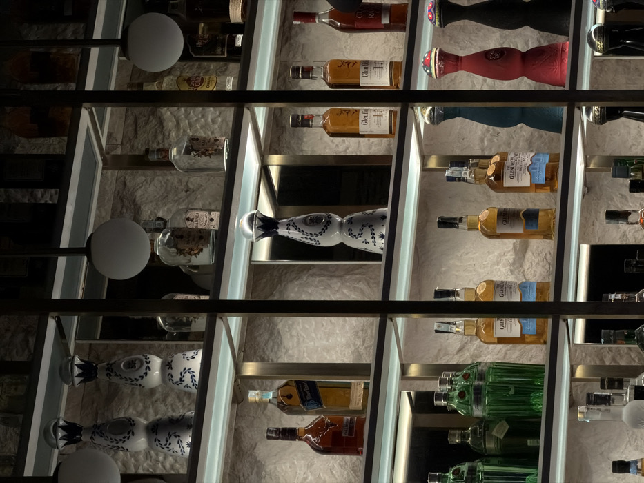 The image displays a multi-tiered shelving unit showcasing various liquor bottles, situated in a bar in Budva, Montenegro. The shelving consists of dark metal frames supporting illuminated clear panels, with each shelf casting light upwards onto the bottles. The background behind the shelves is a textured, light-colored stone wall.
Numerous bottles of different shapes, sizes, and colors are precisely arranged on the shelves. Prominently featured are several tall, white and blue ceramic Clase Azul Tequila bottles, identifiable by their distinctive shape and traditional patterns. Also visible are multiple Glenfiddich single malt Scotch whisky bottles, a Johnnie Walker Blue Label Scotch whisky bottle, and several green-tinted bottles resembling gin. Other brown and clear glass bottles containing various spirits are interspersed. On the lower right, there are several uniquely shaped decorative bottles in solid colors such as blue, red, and black, topped with intricate designs. Two white spherical pendant lights are suspended from above, visible in the upper sections of the frame. No individuals, actions, or interactions are present within the scene. The environment indicates a modern, upscale interior setting, likely a bar or lounge, characterized by accent lighting and a minimalist design aesthetic.