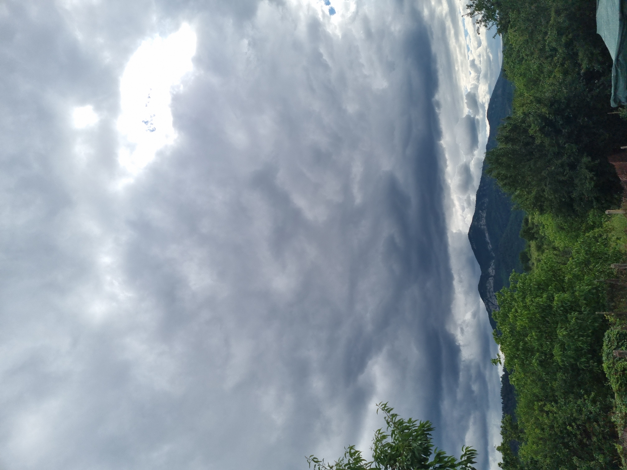 The image shows a natural landscape with a mountain range in the background and lush green trees in the foreground. The sky is filled with dramatic, dark clouds, suggesting an overcast or stormy day. There are some brighter patches in the sky, indicating breaks in the clouds where sunlight is trying to penetrate. The lower part of the image reveals dense vegetation, including trees and bushes, some of which are on the left side of the frame. In the far distance, the silhouettes of hills and the mountain range are visible against the cloudy sky. The scene appears to be taken outdoors in a rural or natural setting, possibly in Dabravka, Bulgaria, given the context. There are no people or animals visible in the image.