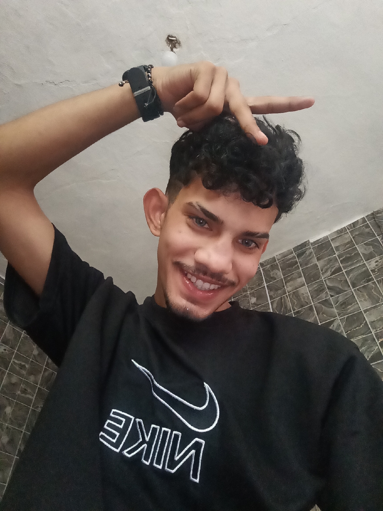 A young man with curly dark hair and light blue eyes smiles at the camera, holding his right hand up to his forehead with his index finger extended. He wears a black t-shirt with a white Nike logo. The background consists of a tiled wall and a white ceiling with a light fixture. The image appears to be a selfie taken indoors.