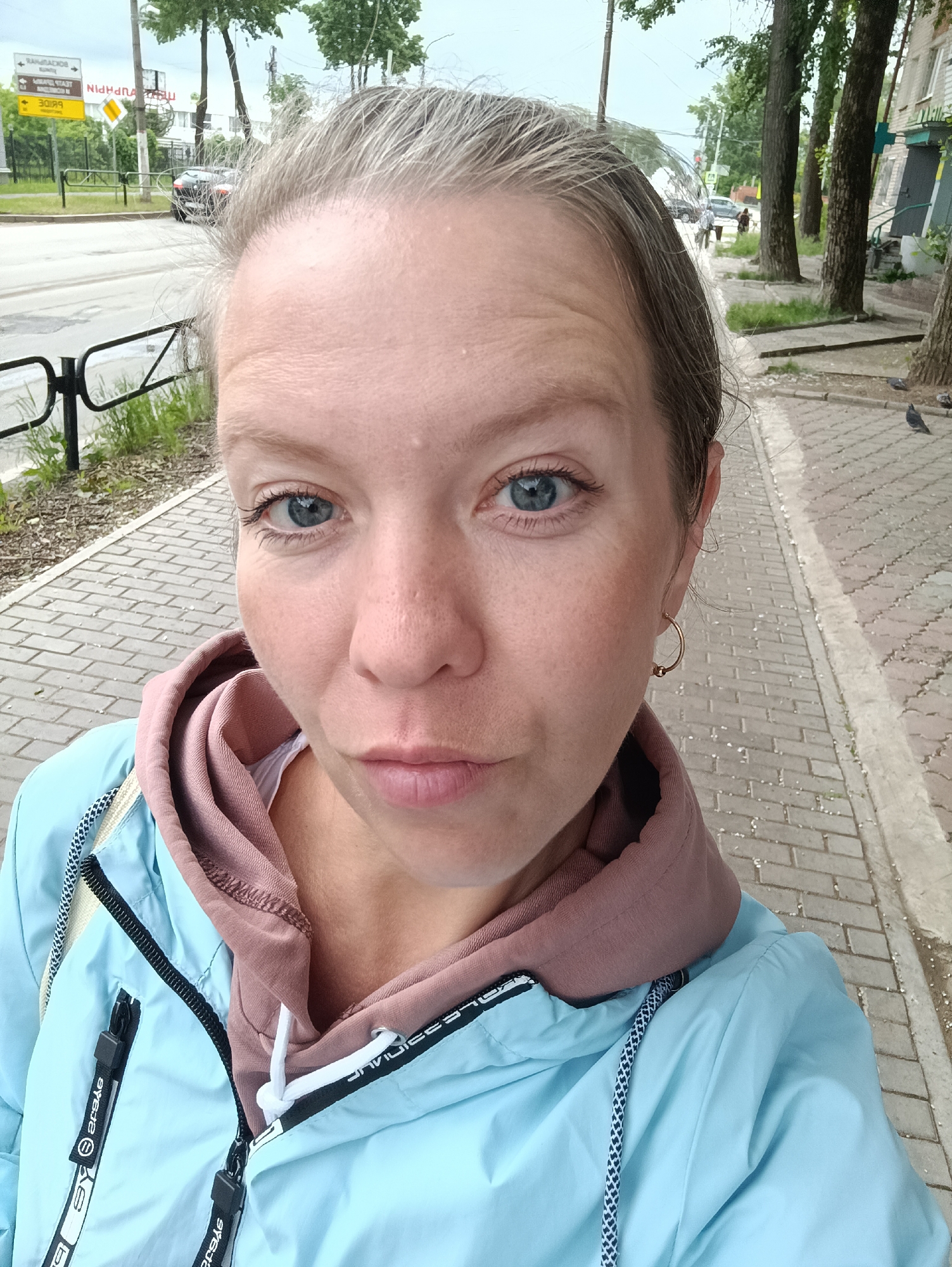 The image is a selfie taken by a light-skinned woman in an urban setting identified as Chaikovsky, Russia. The woman, with light blonde hair exhibiting some graying, looks directly at the camera with blue eyes and a slightly pouting expression. She is wearing a light blue zippered windbreaker over a mauve hooded sweatshirt and a white garment underneath. A small gold-colored hoop earring is visible in her right ear.
The background shows a paved sidewalk to her right, constructed from rectangular gray blocks, and an asphalt road to her left. A black metal barrier separates the sidewalk from the road. On the left side of the road, a pole supports two yellow rectangular road signs. The upper sign displays Cyrillic text including "РАНДРАДИС" (RANDRADIS) and "ДОМ БЫТА" (House of Services), accompanied by a left-pointing arrow. The lower sign exhibits mirrored Cyrillic letters "НИИНАЧАП," which is a reversed form of "ЧАЙКОВСКИЙ" (Chaikovsky), alongside an inverted red-on-white triangular warning symbol, confirming the location as Chaikovsky, Russia. A dark-colored vehicle is partially visible on the road in the distance.
To the woman's right, a strip of ground with sparse grass runs between the sidewalk and several tall, mature trees with rough bark. Beyond these trees, multi-story buildings are visible, one featuring a blue cross symbol, typically associated with a pharmacy ("АПТЕКА"). Several pigeons are present on the sidewalk near the trees. In the far background, indistinct human figures can be seen walking on a distant sidewalk. The sky is overcast.