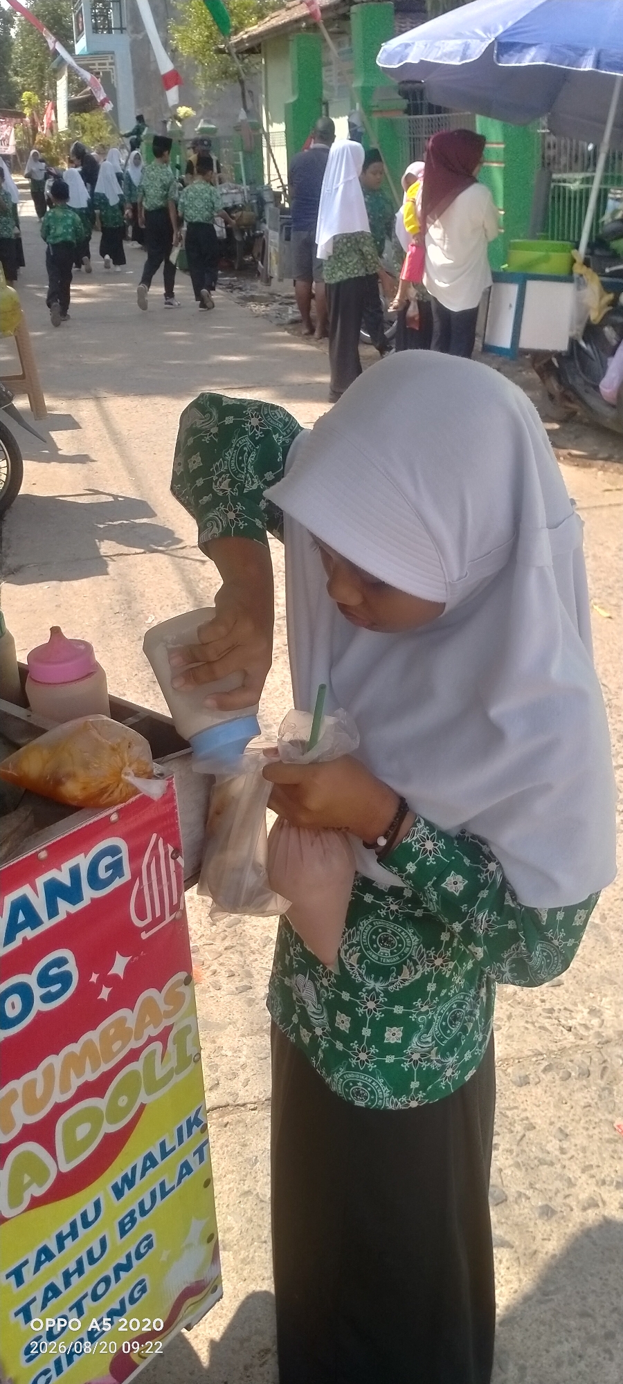 In the city of Sowan Kidul, Indonesia, a young person wearing a white hijab and a green patterned uniform top is dispensing a beverage into a plastic bag. The person is standing next to a cart displaying a sign with text in Indonesian, including "ANG OS TUMBAS ADOLI" and "TAHU WALIK TAHU BULAT TATONG CIRENG." The cart is also holding several plastic bags filled with food items and a pink-capped bottle. In the background, other individuals in similar uniforms are walking down a street lined with buildings, and a blue umbrella is visible. The scene appears to be outdoors on a sunny day.