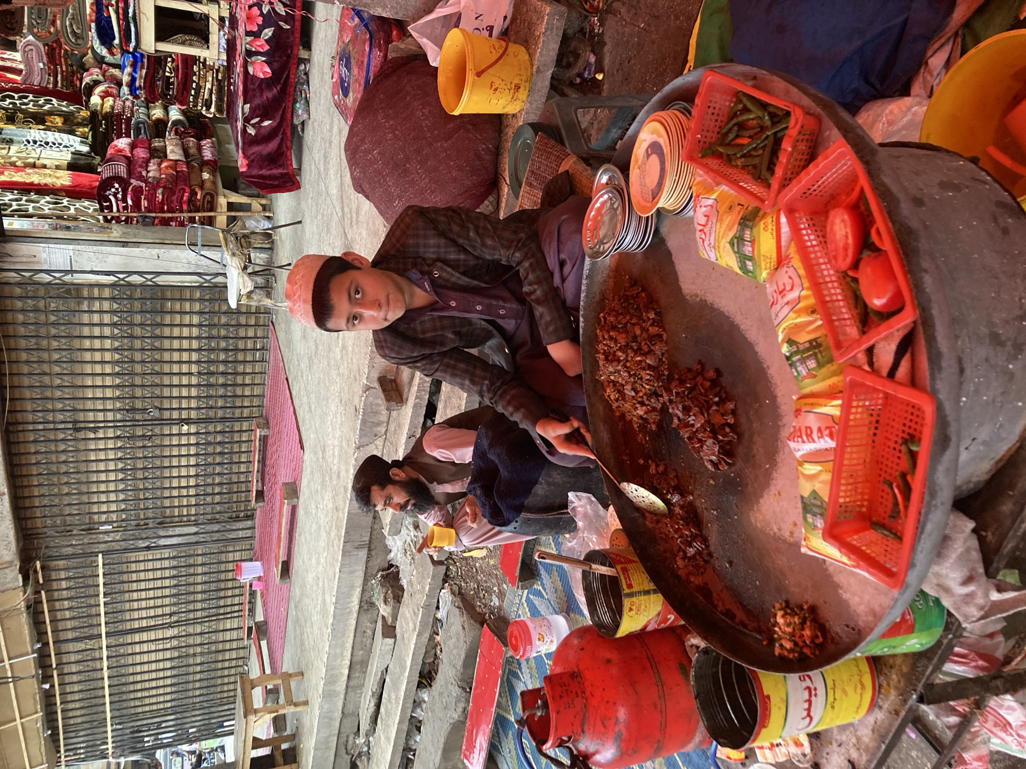 In Quetta City Tehsil, Pakistan, an open-air market stall is depicted on a concrete surface. A young male, approximately 12-16 years old, wearing a light pink skullcap, a dark checkered blazer, and dark trousers, is seated cross-legged in the foreground, looking directly at the viewer. He is positioned behind a large, circular metal griddle containing cooked meat or offal, with a metal slotted spoon resting on the food.
Adjacent to the griddle are stacks of white and patterned serving plates, multiple red plastic baskets holding green chilies, whole tomatoes, and a halved tomato. Packaged food items with Urdu script are also on display. To the young male's left, a large red propane gas cylinder sits on a blue patterned mat, next to a yellow metal can and a black bucket. To his right, a yellow plastic bucket is on the ground.
In the mid-ground to the left, an adult male with a beard, wearing a dark skullcap, light pink shirt, and dark waistcoat, is seated on the same blue mat. He holds a yellow mug in his right hand and appears to be observing something off-frame to the left.
The background features several storefronts. To the left, shops are closed, indicated by their lowered metal accordion security gates. A raised concrete platform in front of these shops holds a light wooden table and several low red seating platforms. To the right, a shop displays numerous stacks of colorful blankets and quilts for sale. The ground beneath and around the stall is concrete, showing signs of wear and scattered debris. An overhead structure, likely an awning or building overhang, is visible at the top of the frame.