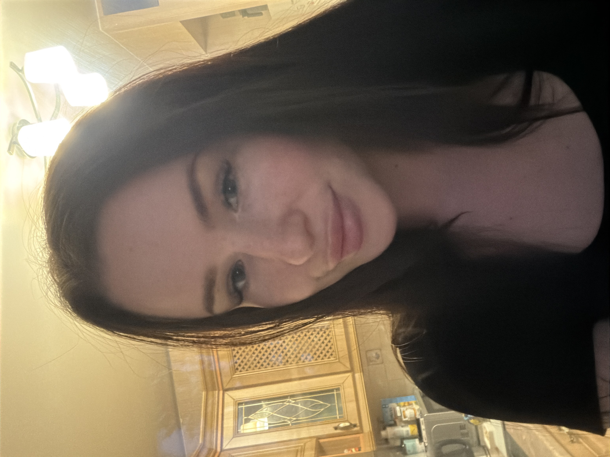 A self-portrait captured in a residential kitchen in Saint Petersburg, Russia, features a woman positioned centrally. She has long, dark hair, dark defined eyebrows, and full lips with a glossy finish. Her gaze is directed towards the camera, and she exhibits a slight smile. She is wearing a dark, round-neck top. The background reveals light-toned wooden kitchen cabinets. The left side displays a cabinet with a glass insert featuring an ornate, curvilinear design, adjacent to another cabinet with a lattice-patterned insert. A countertop with various containers and a silver appliance is visible below the cabinets. An overhead ceiling light fixture with at least two illuminated white cylindrical shades provides ambient lighting, casting a warm hue on the ceiling and upper walls.