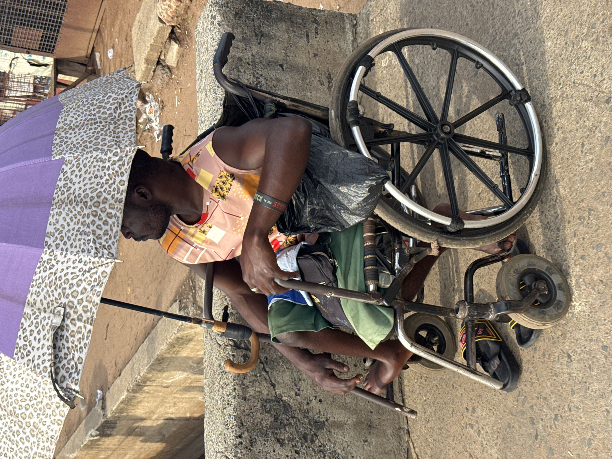 This eye-level, full shot depicts an adult man seated in a manual wheelchair on a sunny outdoor day, likely in Onitsha, Nigeria. The man, with dark skin, is seen in profile looking slightly downwards and to the left. He wears a light-colored, sleeveless top with a geometric or floral pattern in shades of pink, yellow, and orange, along with green shorts. On his right wrist, he wears a dark wristband with faint white text, which appears to read "BACK to the SEMA" or "BACK to the SEMP."
He holds an open umbrella with his left hand, providing shade from the bright sun. The umbrella features alternating purple and leopard-print panels. Both of his hands are engaged with a stick or a part of the wheelchair frame, suggesting he is either propelling himself or manipulating the chair. Another stick with a curved wooden handle rests near his right leg. A black plastic bag is draped over the back of his wheelchair, and a small dark pouch hangs from its frame. His bare feet are visible, one wearing a black flip-flop with yellow accents.
The wheelchair has large black main wheels with silver rims and smaller front caster wheels. It appears sturdy and functional, though showing signs of use. The setting is an urban environment with a concrete or paved ground surface under the wheelchair. To the left, a reddish-brown unpaved area is visible with a large concrete block and some debris. Further in the background, a metal grate structure and other indistinct elements of a street or marketplace can be seen. The bright sunlight indicates it is daytime, casting clear shadows.