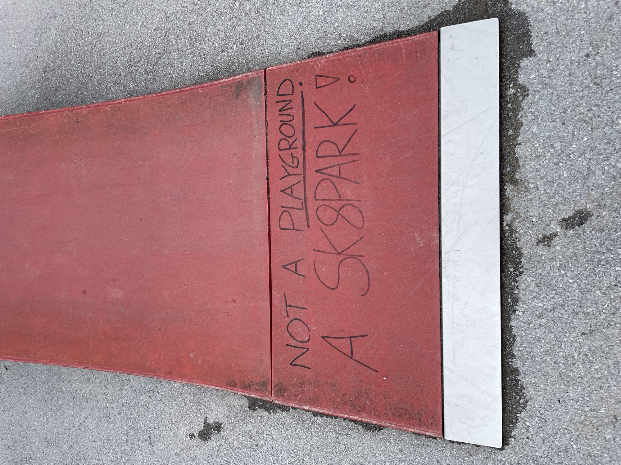 The image shows a red, curved concrete structure placed on asphalt in Unknown, Unknown. Handwritten in black marker on the concrete is the phrase "NOT A PLAYGROUND A SK8PARK!". The end of the concrete structure is bordered by a white concrete strip.