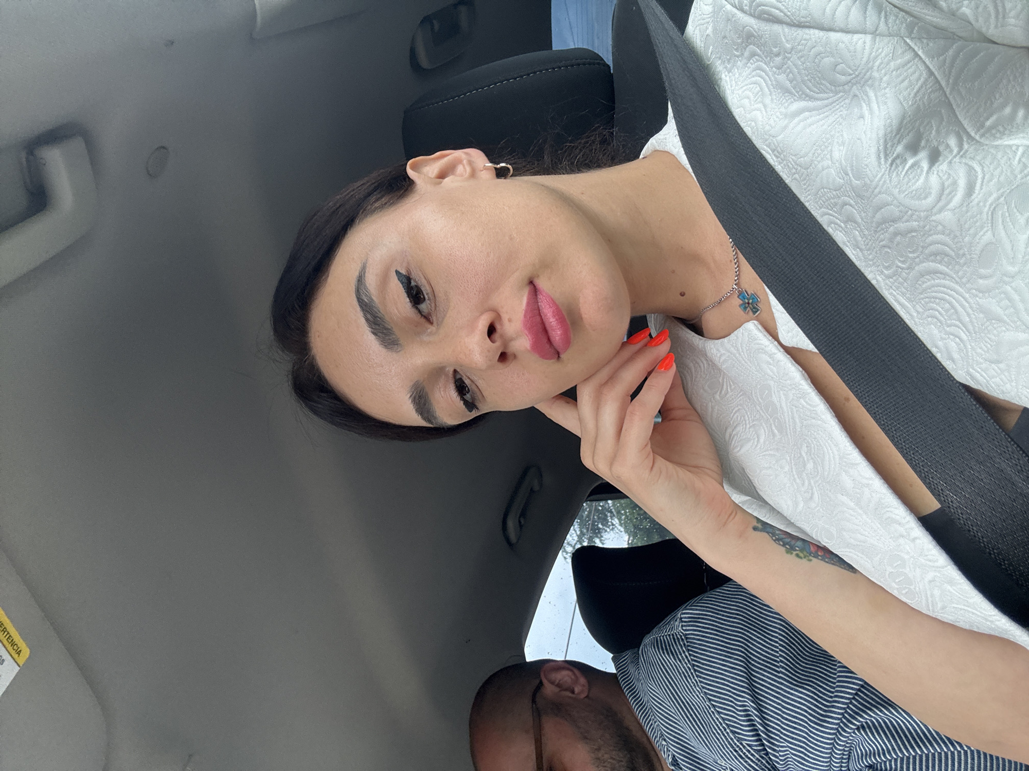 A woman is taking a selfie in the passenger seat of a car, with a man sitting in the driver's seat. The woman has dark hair and is wearing a white jacket over a dark top. She has bright pink lipstick and her nails are painted bright orange. She is wearing a silver necklace with a blue pendant. The man has a shaved head and is wearing glasses and a striped shirt. The car appears to be moving, as indicated by the slight blur in the background and the overall composition of the image. The interior of the car is visible, including the dashboard and ceiling. There is a yellow sticker on the car's ceiling, which appears to be a warning label. The background visible through the car windows suggests a location with trees and buildings, indicating an urban or suburban environment. The image was taken in Tbilisi, Georgia.