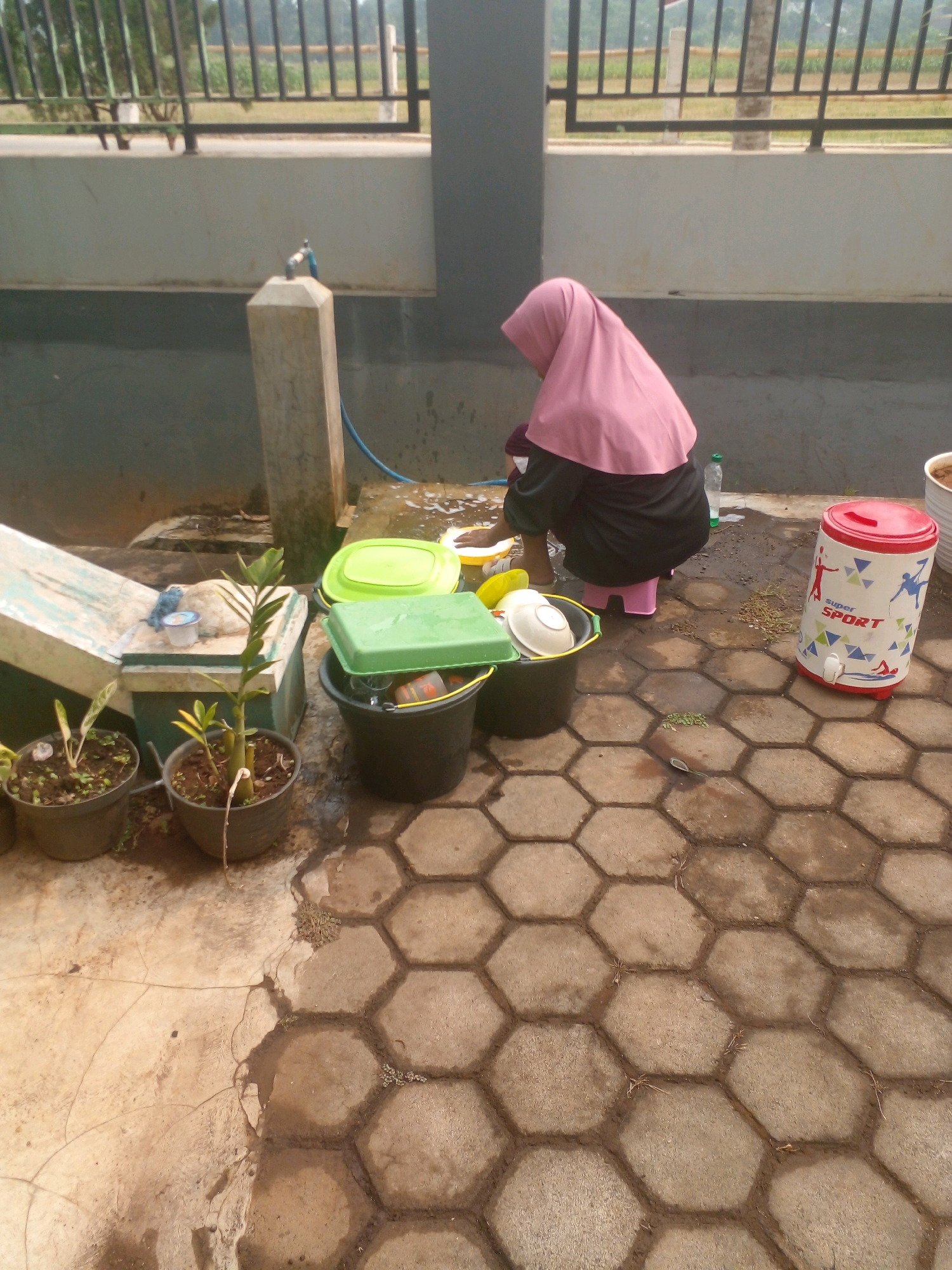 In Banyuputih, Indonesia, a woman wearing a pink hijab and a dark abaya is crouching on a pink stool to wash dishes outdoors. She is positioned to the right of a large black bucket containing various dishes and green lids. To her left are two potted plants. Behind her, a water faucet extends from a concrete pillar, with a blue hose leading away. Further back, a dark grey wall with a lighter grey trim runs horizontally across the scene, topped by a black metal fence. The ground is paved with hexagonal concrete tiles, some of which are wet. A red and white cylindrical water cooler with the "Super Sport" logo sits to the right of the woman.